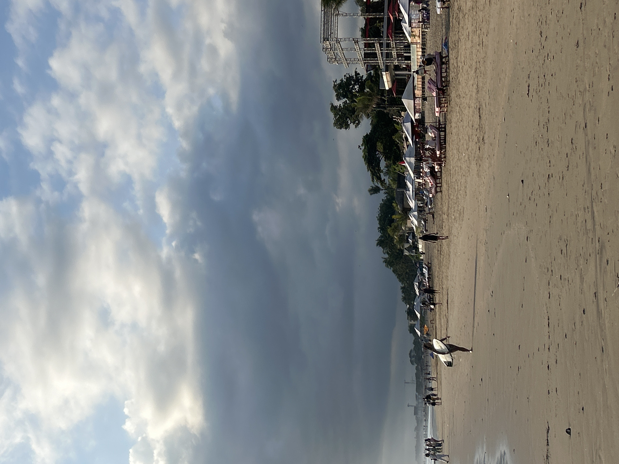 The image depicts a wide beach in North Kuta, Indonesia, with overcast skies casting a diffused light. In the foreground, a person carries a surfboard away from the water. The beach is lined with numerous lounge chairs and umbrellas, some occupied by people. A few more individuals are walking along the shore. In the background, lush palm trees and a multi-story structure with scaffolding are visible, suggesting a developed resort or entertainment area. The overall scene conveys a relaxed atmosphere on a cloudy day at a popular beach destination.