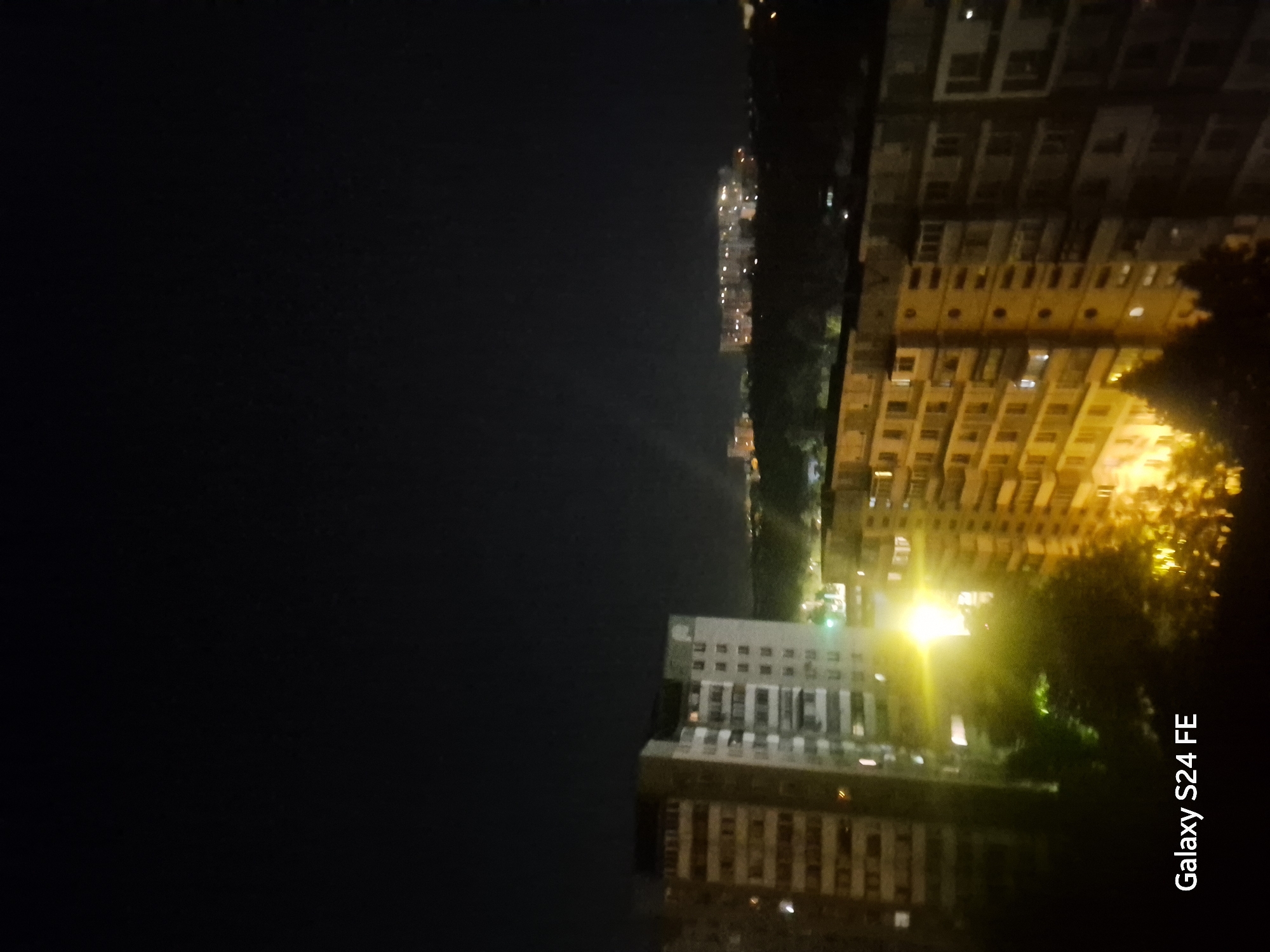 The image is a nighttime aerial view of a residential area in Chișinău, Moldova. In the foreground, two large apartment buildings are visible. The building on the left is a multi-story structure with many windows, some of which are illuminated. To its right, another similar building is partially visible. In the middle ground, trees and smaller buildings with illuminated windows can be seen. The background features more distant buildings and the dark expanse of the night sky. The bottom left corner of the image displays the text "Galaxy S24 FE". The scene is illuminated by artificial lights from the buildings and streetlights, creating a stark contrast with the dark sky.