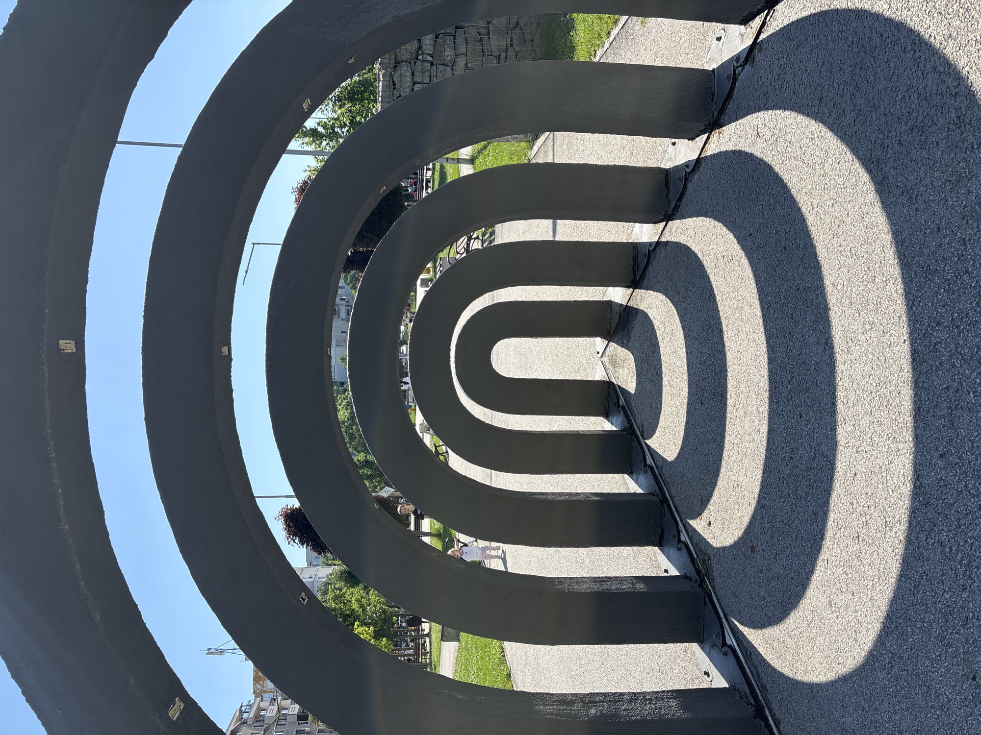 The image shows a public art installation in Plovdiv, Bulgaria, consisting of concentric, arch-shaped metal structures that create a tunnel-like effect. The arches are painted a dark gray and are spaced closely together. The ground beneath the arches is paved. Shadows cast by the arches form a repeating pattern of curved lines on the ground. Beyond the installation, a green lawn and trees are visible. In the background, buildings and a street lamp can be seen. Two people can be seen between the arches.