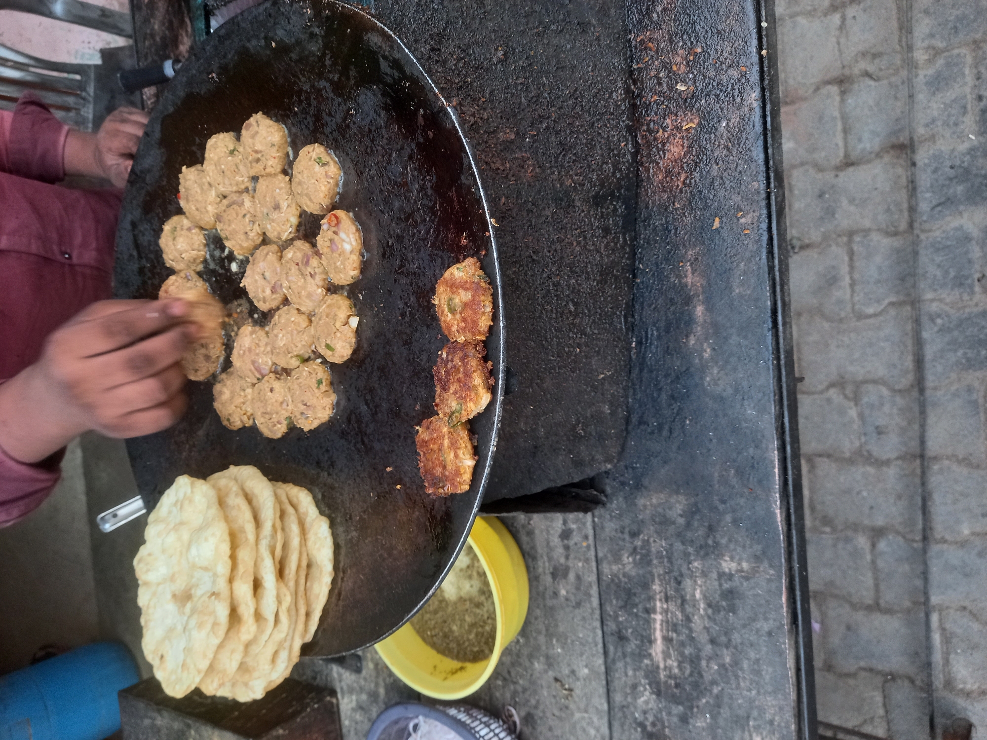 A street vendor in Kalpi, India prepares food on a large griddle. The vendor, wearing a maroon shirt, arranges small, round patties on the griddle with his hands. Several cooked patties are visible on the right side of the griddle. A stack of unleavened bread sits on the edge of the griddle. A yellow container holding spices or other ingredients is visible beneath the griddle. The scene appears to be set up on a street or walkway, judging by the paved area to the side of the griddle. The entire scene is viewed from directly overhead.