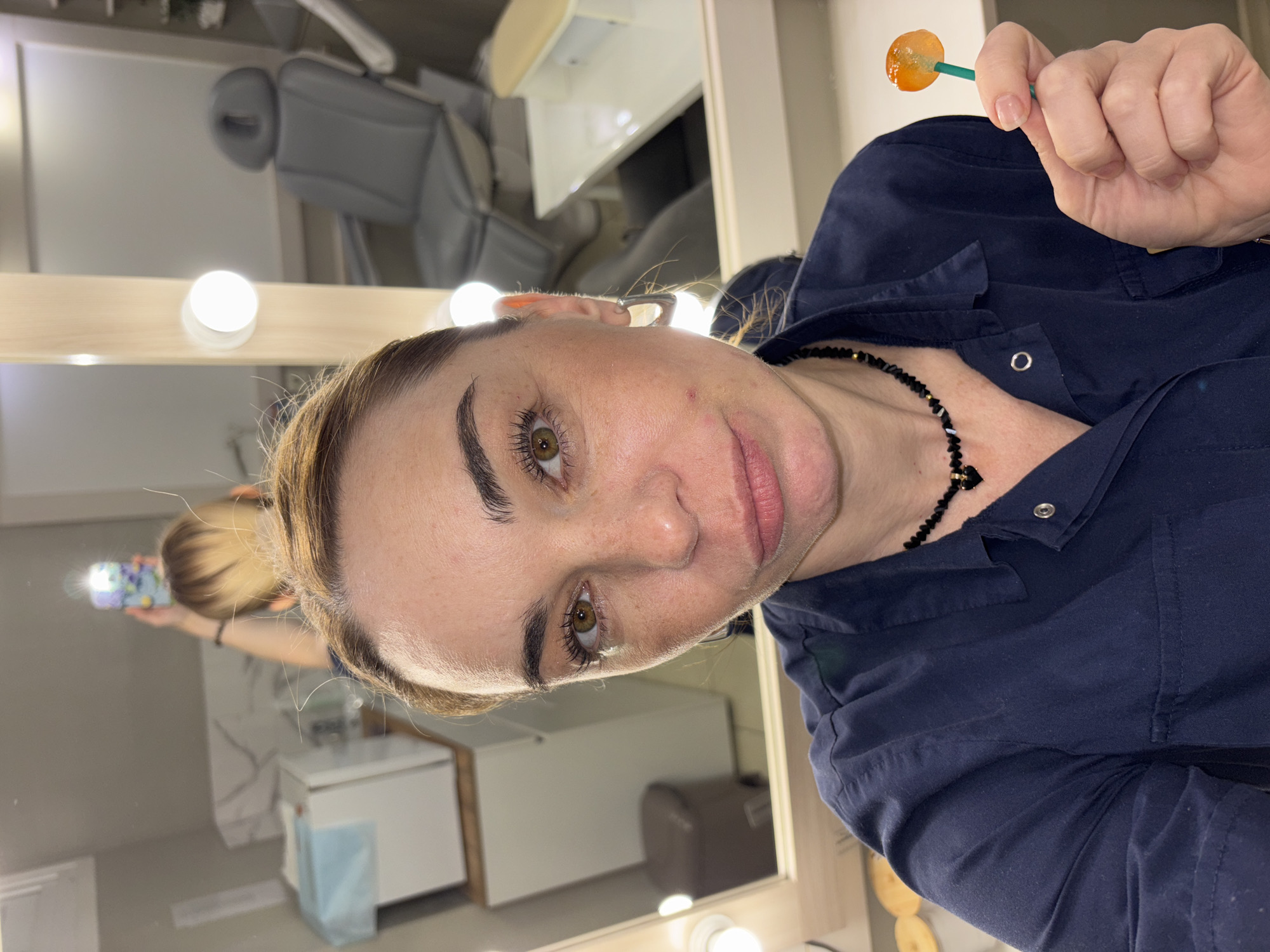 A woman with blonde hair pulled back in a bun is holding an orange lollipop. She is wearing a dark blue shirt and a black beaded necklace. She appears to be in a room with a vanity mirror surrounded by lights. A reflection of a person holding a phone can be seen in the mirror. Other objects in the reflection include medical equipment, likely for cosmetic procedures, possibly indicating the woman is in a clinic or salon. There is no indication that the scene is in Vsevolozhsk, Russia.