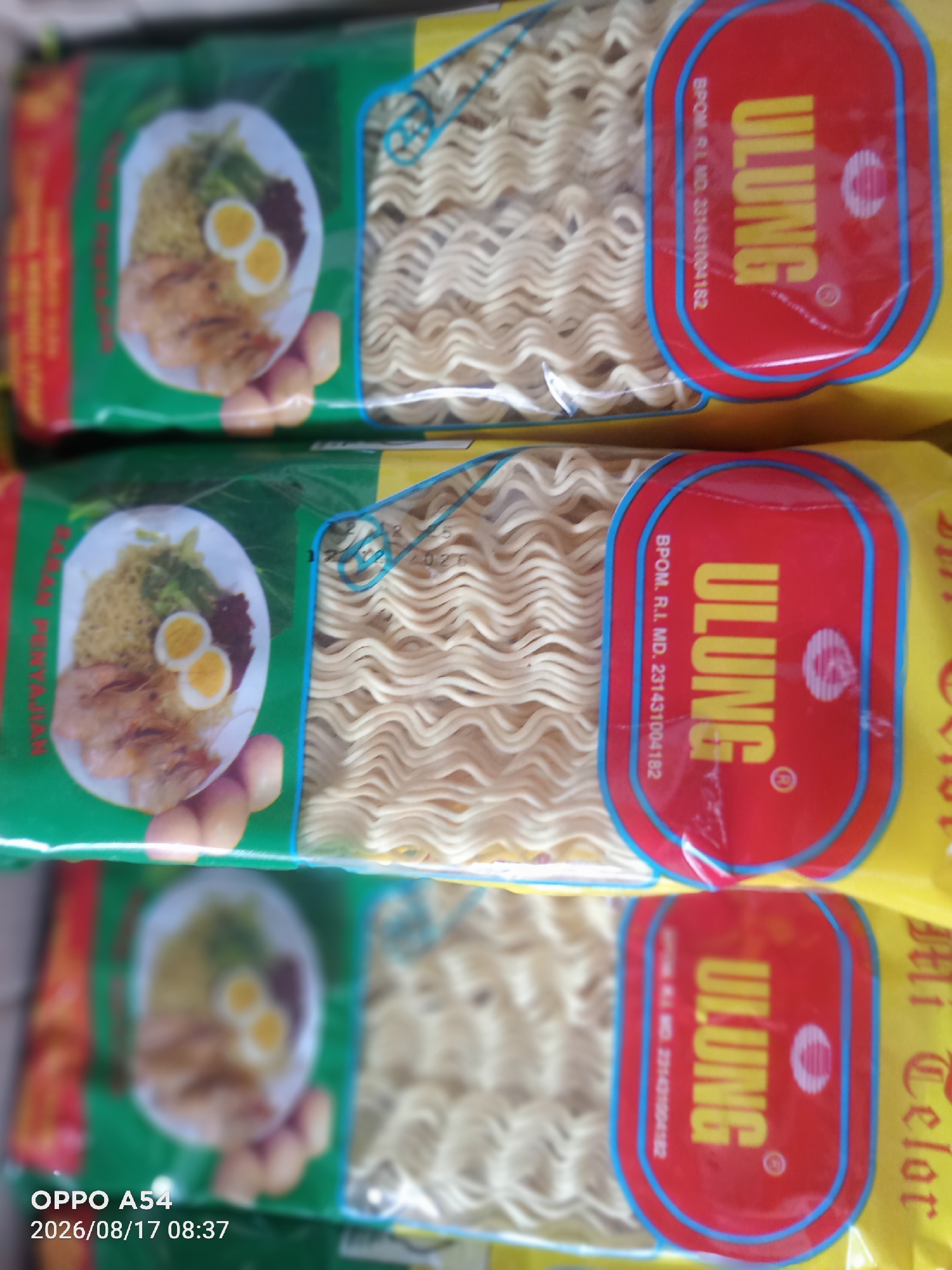 The image displays three packages of ULUNG instant noodles stacked vertically. Each package features a green and yellow design with a red oval logo containing the brand name "ULUNG" in yellow lettering. The logo also includes a circular symbol and text in a smaller font, "BPOM. R.I. MD. 231431004182." To the left of the logo on each package is a blurred image of a serving suggestion, showing noodles, two halves of a boiled egg, and what appears to be a protein. The noodles themselves are visible within clear plastic packaging. The bottom left corner of the image shows a timestamp: "OPPO A54 2026/08/17 08:37." The background is out of focus, suggesting the packages are likely on display or in storage. There are no people visible in the image. The scene is consistent with a retail environment, potentially a market or grocery store in Jambi, Indonesia, where this product is sold.
