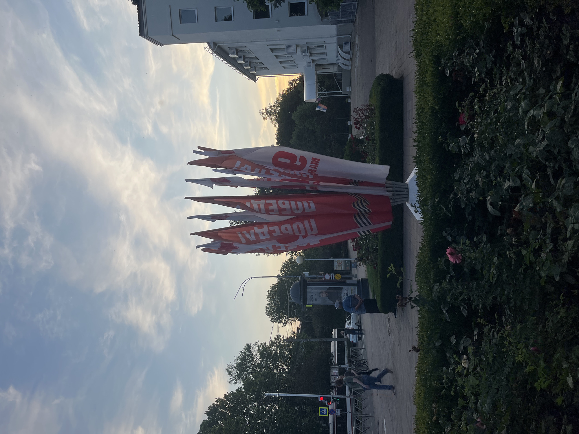 The image depicts a street scene in Krasnodar, Russia, likely during the late afternoon or early evening, given the lighting and sky conditions. In the foreground, a cluster of red and white flags, many bearing the word "ПОБЕДА" (Victory) and images of the Order of the Patriotic War, are prominently displayed. These flags are mounted on a central pole structure and appear to be part of a commemorative display.
To the right, a modern white building with multiple windows and air conditioning units is visible. The building's facade is clean and well-maintained. A small Russian flag is visible near the entrance of this building.
In the middle ground, a sidewalk area is busy with pedestrian activity. People are walking in different directions, and a bicycle is parked near a lamppost. A public transportation vehicle, possibly a trolleybus or tram, is stopped at a traffic signal, which is red. A street advertisement column is also present, featuring a portrait of a man. Trees line the street, creating a leafy backdrop.
The environment is urban, with paved sidewalks and roads. Landscaped green areas with hedges and rose bushes are visible in the foreground and middle ground, suggesting a well-maintained public space. The sky above is partly cloudy with streaks of cirrus clouds, illuminated by the setting sun, which casts a warm glow on the scene.