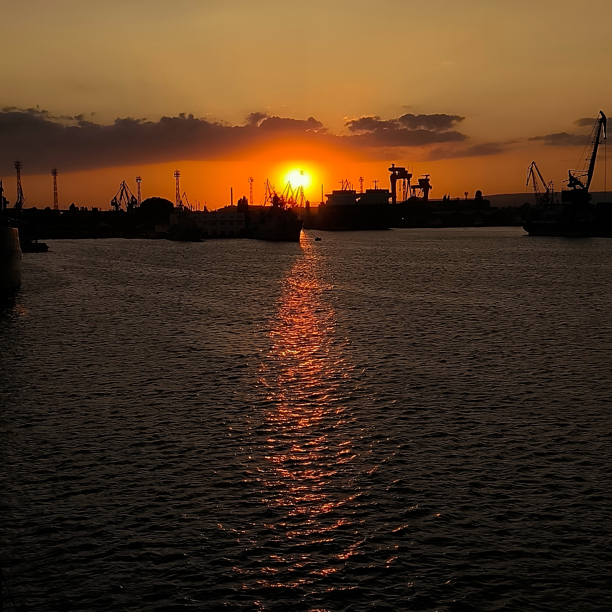 This is a vertical, eye-level shot of a harbor at sunset in Varna, Bulgaria. The sky is filled with a warm orange glow, with darker clouds towards the top. The sun is a bright, fiery orb, partially obscured by clouds, casting a shimmering reflection on the dark, rippling water. The silhouette of a cityscape, featuring various cranes, ships, and buildings, lines the horizon. The overall atmosphere is serene and industrial.