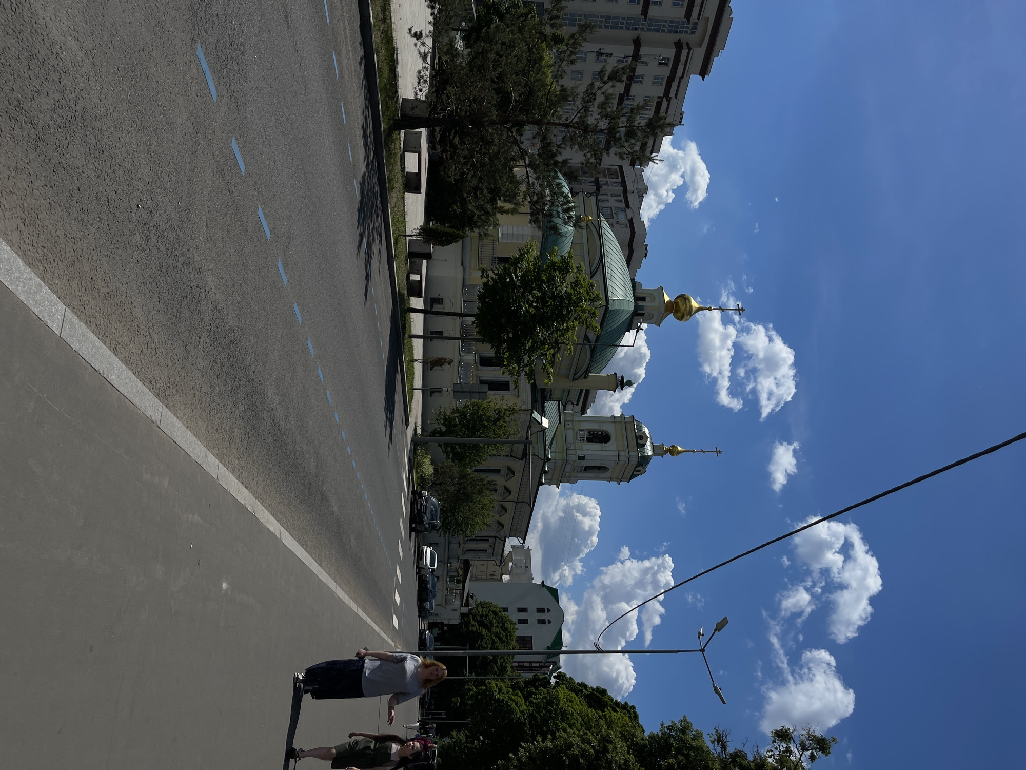 The image captures a vibrant urban street scene in what appears to be Moscow, Russia, under clear, bright daylight. The perspective is from a slightly elevated position looking down a road that runs diagonally from the lower left towards the upper right.
In the foreground, two individuals are standing on a paved surface, possibly a sidewalk or pedestrian area adjacent to the road. One person, closer to the camera on the left, wears a light gray t-shirt and dark shorts or pants. The second person, slightly behind and to the right, wears a dark green or brown t-shirt and shorts. Both cast long, distinct shadows to their left, indicating strong sunlight from the right.
The road itself is asphalt, marked with multiple dashed blue lines. A paved curb separates the road from a grassy area and trees on the right side. In the mid-ground and background, a prominent church or cathedral with light-colored walls, green domes, and golden cupolas topped with crosses stands out. Its architecture suggests a Russian Orthodox style. Various trees are interspersed around the church and along the street, providing greenery. Behind the church, several multi-story modern residential or commercial buildings with numerous windows are visible. A few parked cars are also seen on the street.
The sky is a vivid blue, dotted with fluffy white cumulus clouds. A streetlight with multiple lamps is positioned in the lower right, and a power line stretches across the upper right portion of the sky. The bright conditions and the sharp shadows suggest it is daytime, likely morning or early afternoon. No discernible text is visible in the image.