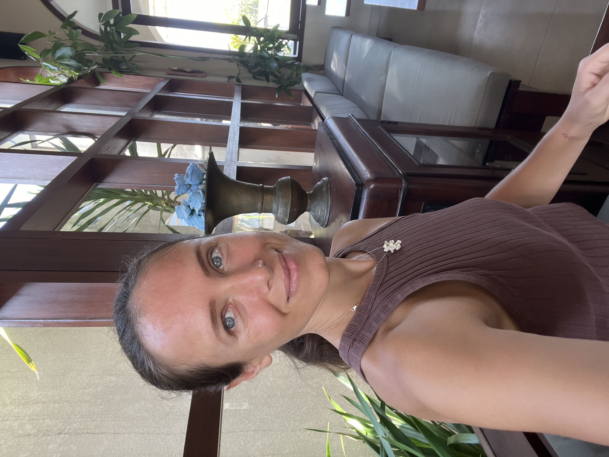 A woman with shoulder-length brown hair and blue eyes smiles at the camera in a well-lit room. She is wearing a brown sleeveless ribbed top and a necklace with a white pendant. The background features a wooden grid structure, plants, and a sofa. A brass vase with blue flowers sits on a wooden cabinet. The scene appears to be indoors, possibly a cafe or lounge area in Ubud District, Indonesia, given the tropical plants and architectural style.