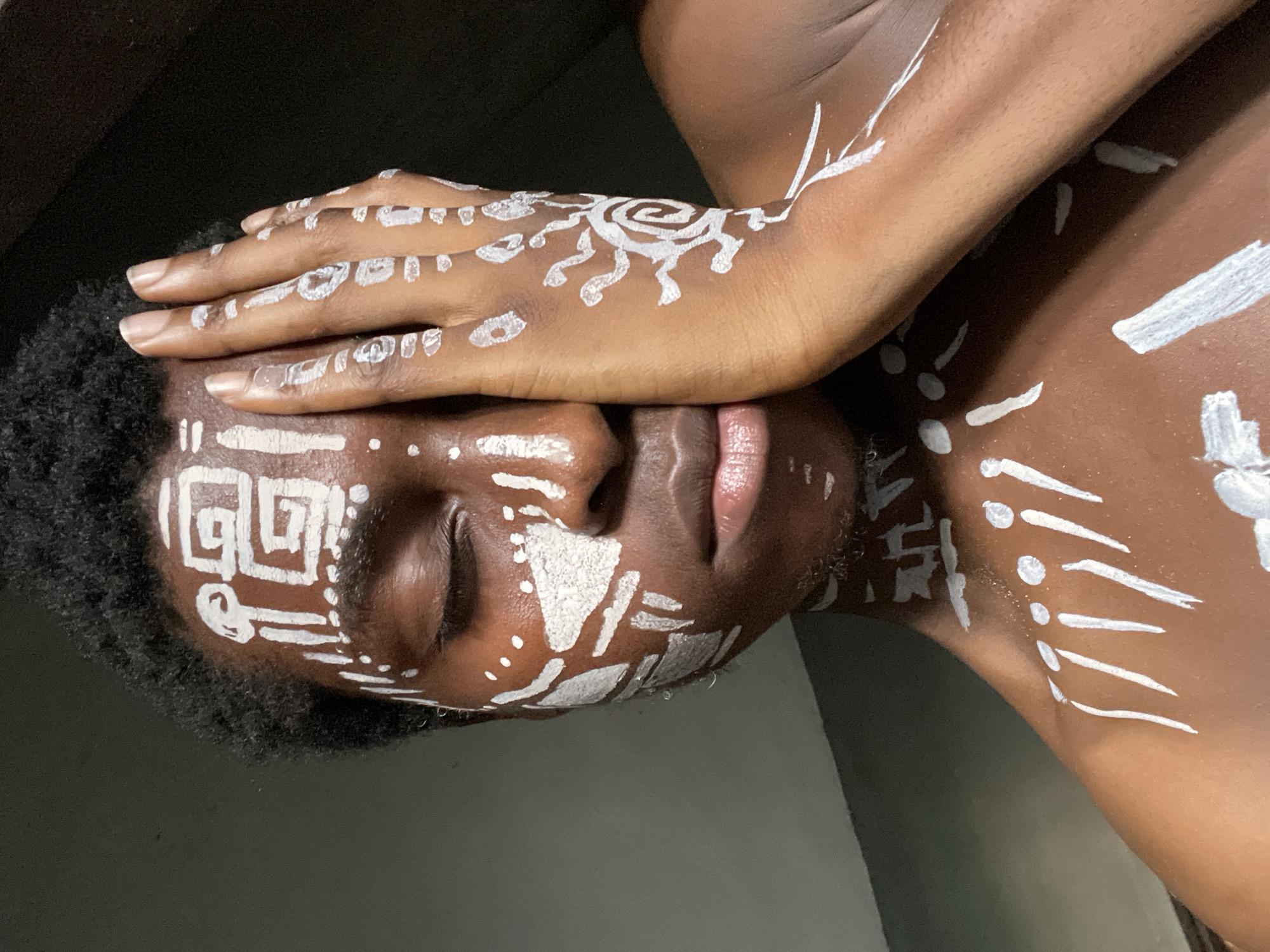 The image portrays a person with dark skin, positioned sideways with their head tilted. One of their hands is placed over their closed eyes and forehead, partially obscuring the upper part of their face. The person has white body paint applied in intricate patterns across their face, hands, and visible upper body.
On the hand covering the face, the paint features small dots and short, abstract lines. A notable design on the forearm is a prominent circular or spiral motif with radiating lines, resembling a stylized sun or a complex labyrinth. The face is adorned with geometric patterns, including a square spiral design on the forehead, various strokes, and dots on the cheeks and chin. Abstract white strokes and dots also extend across the person's chest and shoulder area.
The person's eyes are closed, and their lips are slightly parted. Short, dark curly hair frames the side of their head. The lighting is soft, creating a stark contrast between the dark skin tone and the vivid white paint. The background is uniformly dark, directing focus entirely to the subject. The pose, with closed eyes and a hand covering the face, suggests contemplation, meditation, or a symbolic gesture. While the specific cultural context of the body paint is not explicitly detailed in the image, such practices are prevalent in various cultures, including those in regions like Onitsha, Nigeria. There is no visible text present in the image.