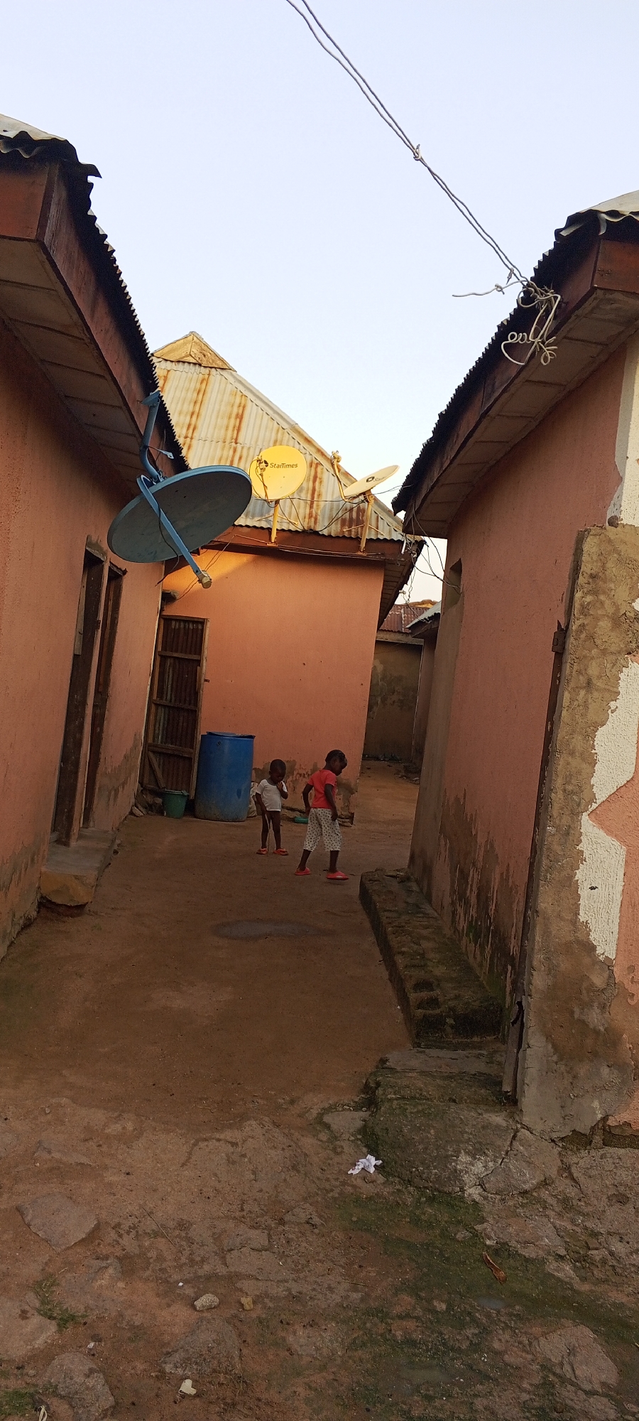 The image shows a narrow alleyway between two buildings with orange-colored walls. Two young children are standing in the middle of the alley, facing away from the camera. The child on the left is wearing a white t-shirt and white shorts, while the child on the right is wearing a red t-shirt and patterned shorts. A large blue barrel is situated against the wall to the left, and a green bucket is next to it. Above the alley, a satellite dish is attached to the side of the building on the left. The ground is dirt and has some scattered rocks and debris. Overhead, power lines are visible against a clear sky. The architectural style of the buildings, with corrugated metal roofs and simple construction, suggests a residential area in Jalingo, Nigeria. The scene depicts a casual outdoor moment for the children in their environment.