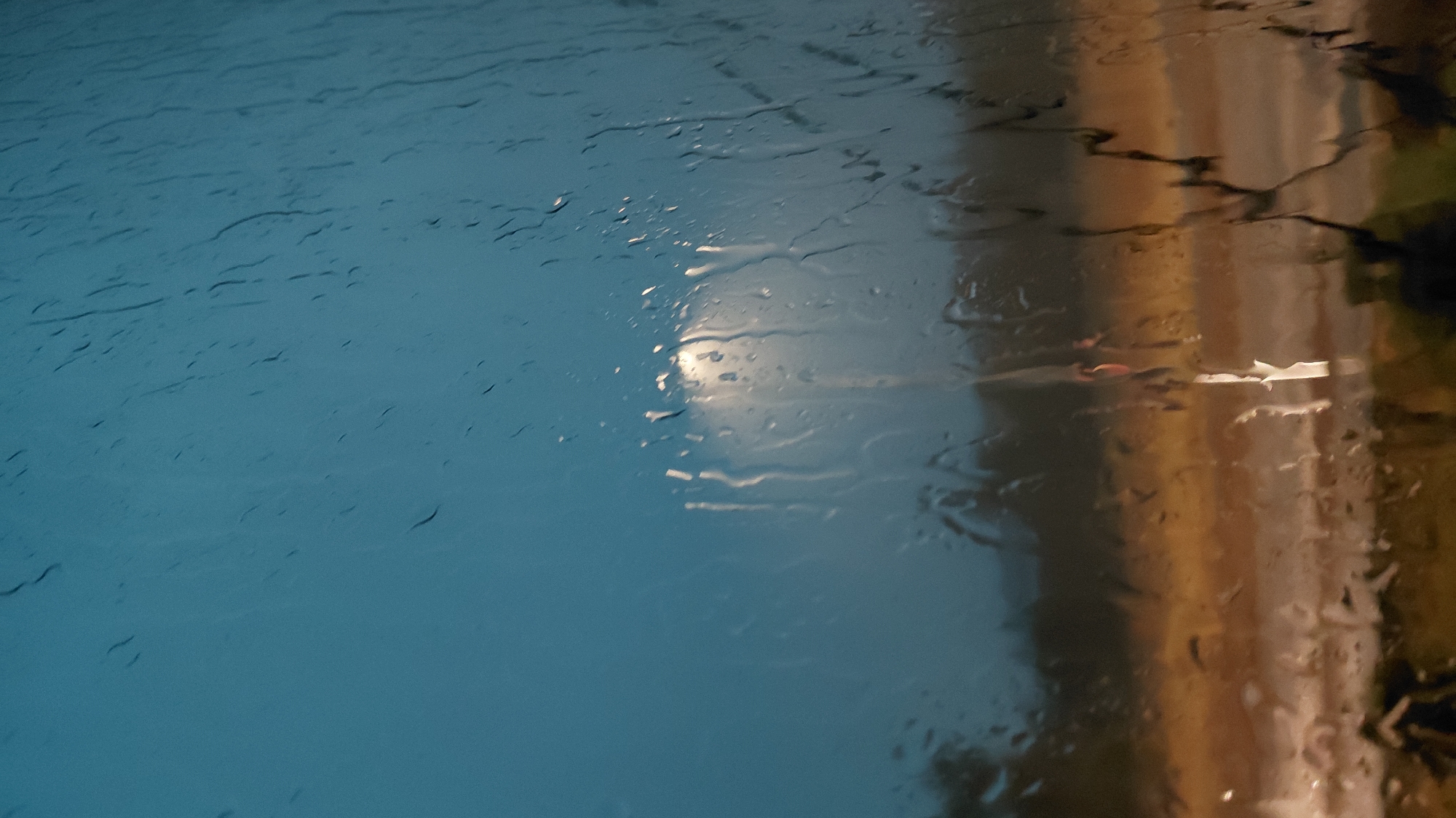 This is a photograph taken through a window covered in raindrops, creating a blurred and distorted view of the environment outside. The sky is a deep, muted blue, suggesting it is twilight or overcast. In the middle ground, a bright, circular light source, likely a streetlamp, is visible, diffused by the raindrops. Below the light, a darker horizontal band suggests trees or buildings, and further down, a lighter, horizontal band of warm, golden light could be a road or illuminated area. The foreground is dominated by the streaks and droplets of water on the glass, obscuring details of any potential subjects or objects. The overall impression is one of looking out at a dimly lit, possibly urban or suburban scene during a rainy period. The location is specified as Abovyan, Armenia.