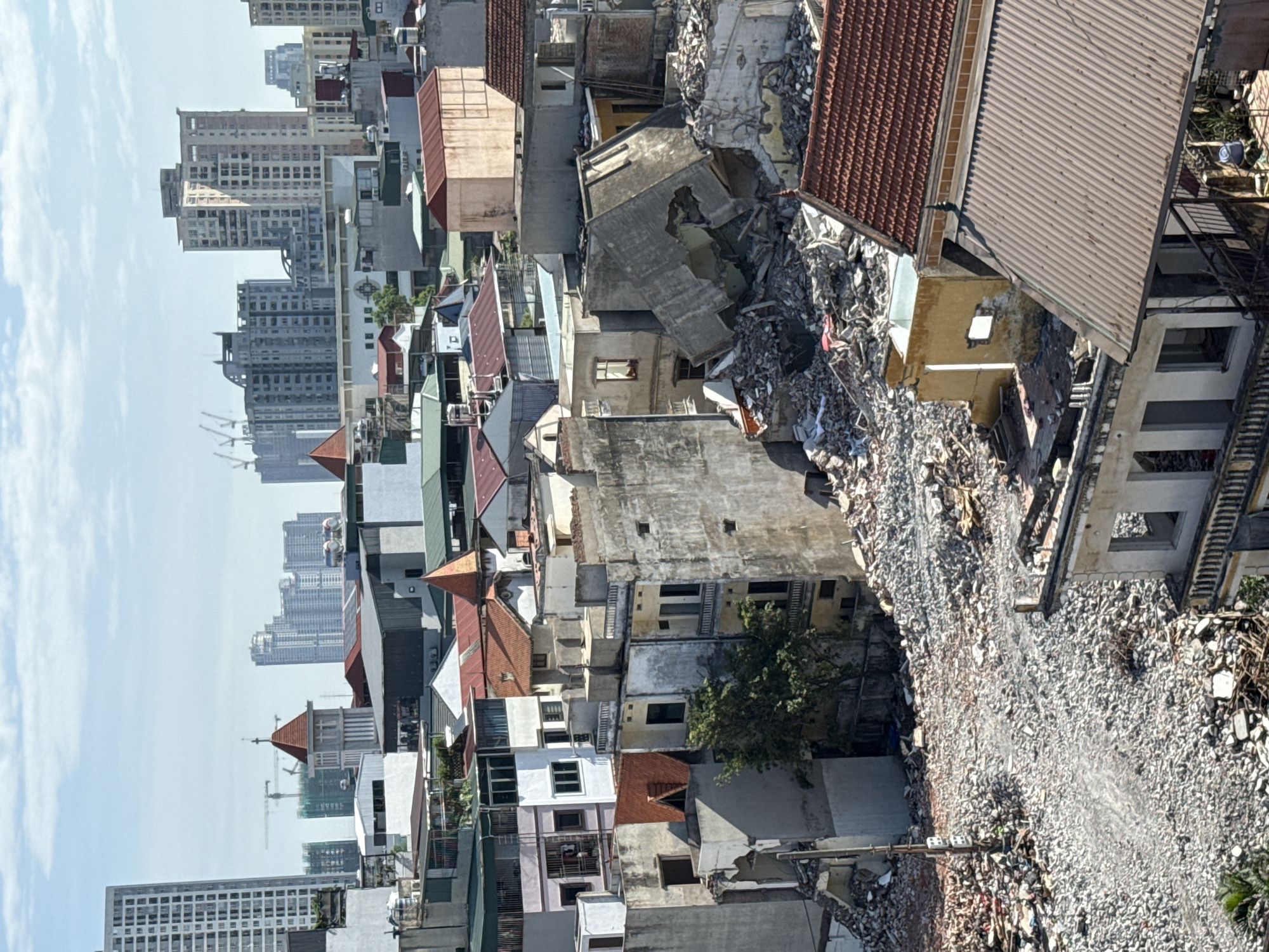 The image shows a dense urban environment in Hà Nội, Vietnam. In the foreground and center, there are old, tightly packed buildings, some damaged or in the process of demolition, creating a path of rubble. The buildings vary in height, style, and color, with many having tiled or metal roofs. A few trees are visible among the structures. In the background, a skyline of modern high-rise buildings is present. The sky is clear and blue.