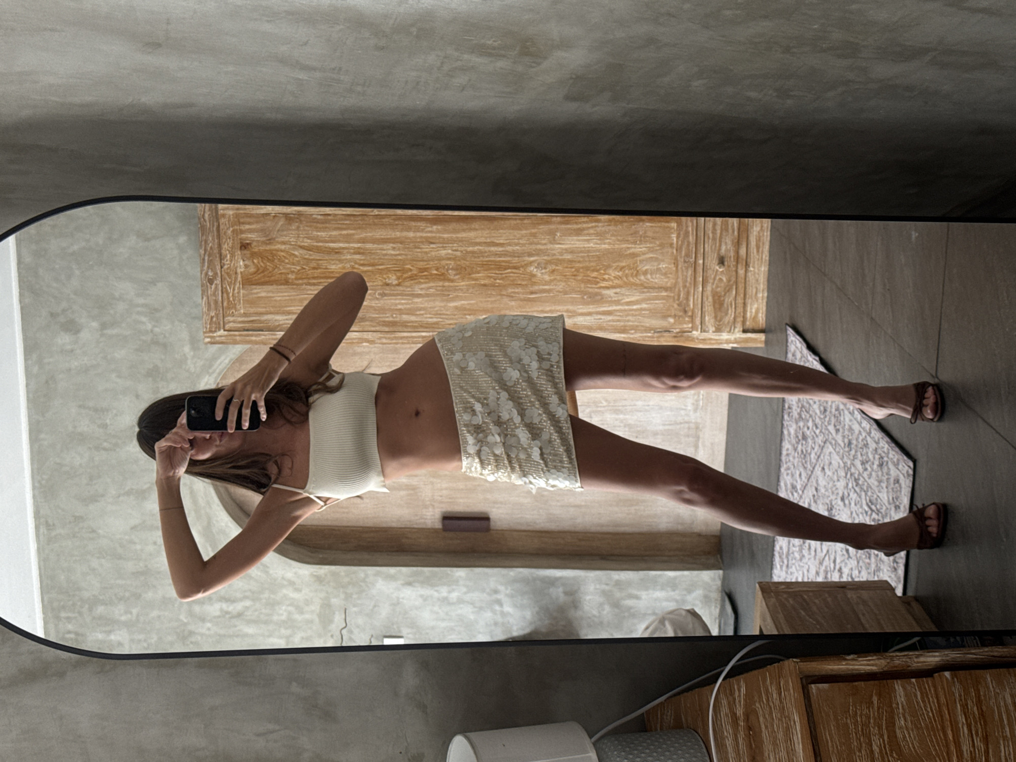 A woman is reflected in a full-length mirror. She is wearing a white crop top and a short, off-white skirt with floral embellishments, along with brown sandals. She is holding a black smartphone up, seemingly taking a photo of herself in the mirror.
The setting appears to be the interior of a room, featuring light gray walls and light wooden furniture. A patterned rug is partially visible on the floor. The location is identified as North Kuta, Indonesia.