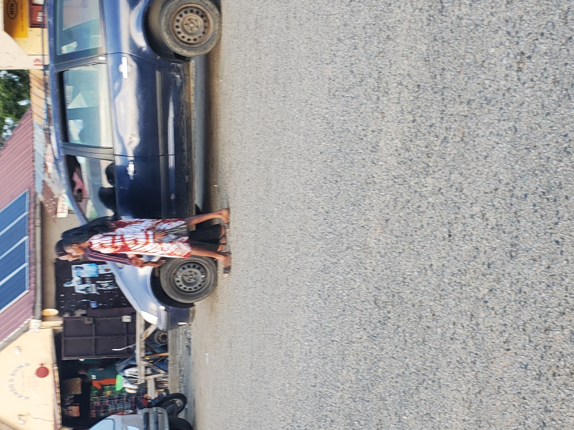 In Ampan, Nigeria, a dark blue station wagon is parked on a gravel road beside a building with solar panels on its roof. Two individuals, one wearing a white and red patterned dress and the other in a maroon top and dark trousers, walk past the car. The building appears to be a shop, with goods visible inside. To the left of the station wagon, a portion of a lighter-colored vehicle and a motorcycle are also visible. The scene suggests a typical roadside commercial area.