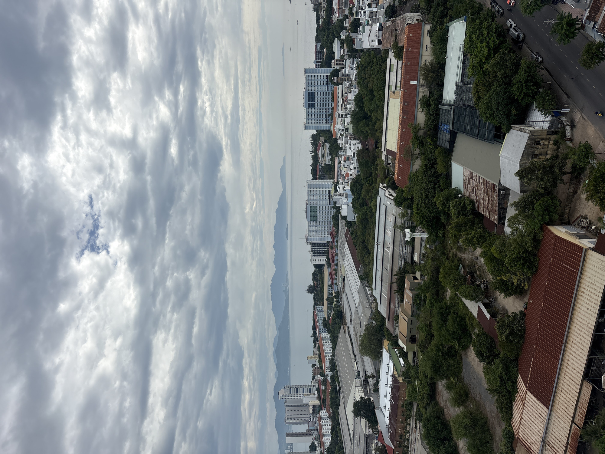 This is an aerial, landscape-oriented photograph taken from a high vantage point, overlooking a coastal city in Nha Trang, Vietnam. The image is dominated by a vast, cloudy sky filling the left two-thirds of the frame, with a mix of dark and light gray clouds stretching horizontally. The right third of the image reveals the city, characterized by a dense urban landscape juxtaposed with lush greenery and a body of water.
The cityscape features a variety of buildings, from tall, modern apartment complexes with white facades and numerous windows to lower-rise structures with red and white tiled roofs, suggesting a mix of residential and commercial or industrial areas. Trees are abundant, growing between buildings and on the slopes of hills that rise behind the city. A road with several cars and at least one motorcycle is visible, indicating active urban life.
In the distance, a calm body of water, likely the sea, separates the city from a range of hazy, blue mountains. The presence of boats on the water hints at maritime activity. The overall atmosphere is overcast, suggesting a cloudy day, possibly in the morning or late afternoon given the diffused light. The scene captures a vibrant, developing urban environment nestled against a natural coastal backdrop. There is no visible text in the image.