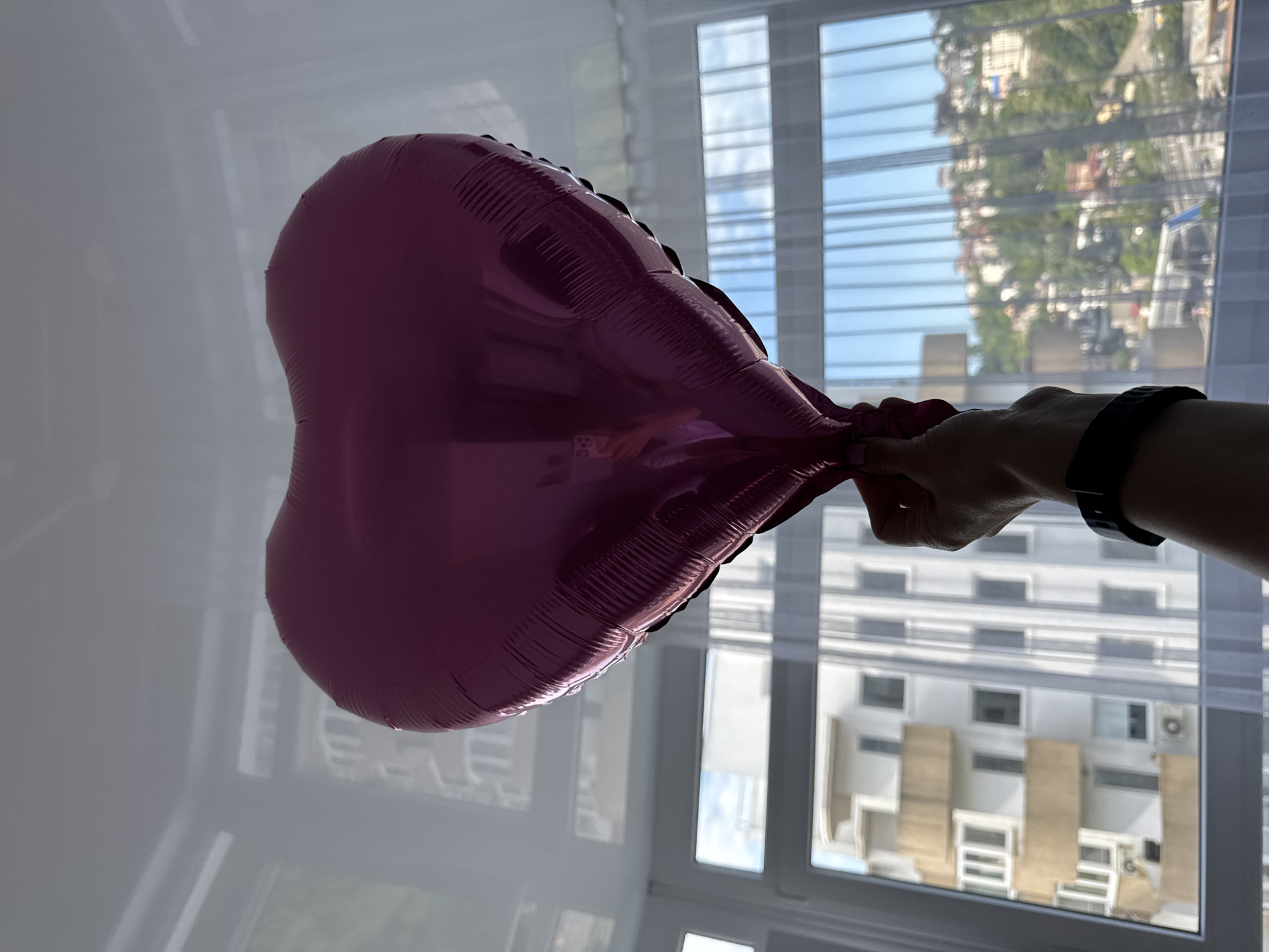 A heart-shaped pink foil balloon is held by a person, silhouetted against a sunlit window. The person's arm and wrist are visible, adorned with a dark watch. Outside the window, the reflection of a building is visible, suggesting the location is Sochi, Russia, given the architectural style and visible foliage. The interior is brightly lit, with a white wall partially visible to the left of the frame. The window has vertical blinds or bars and reflects the cityscape during the daytime.