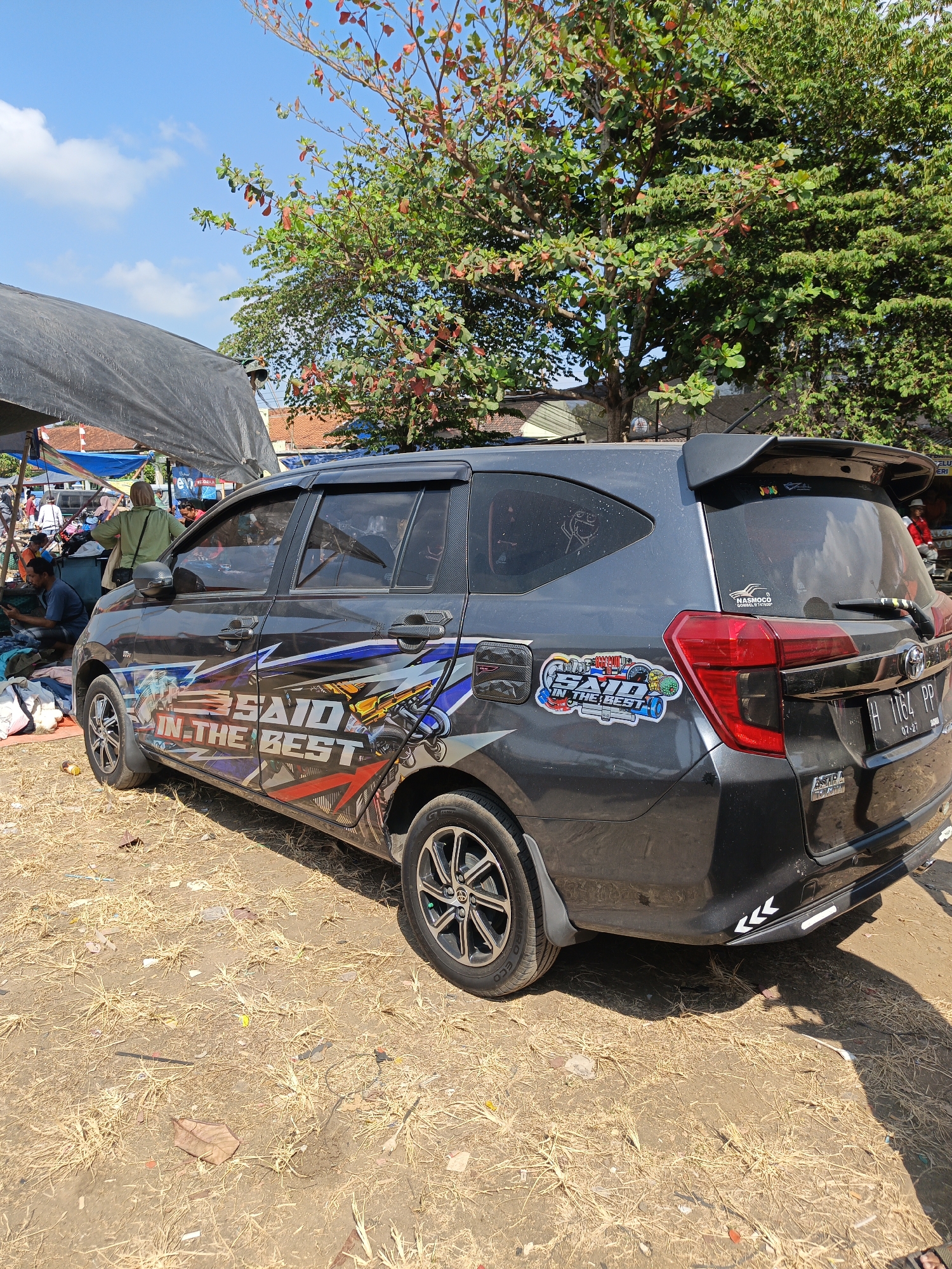 A dark gray minivan is parked on a dirt ground under a clear blue sky with scattered white clouds. Trees with green and red leaves are visible in the background. A large black tarp is partially covering the minivan and the area to its left. Several people are visible under the tarp, some appearing to be selling goods, while others are sitting or standing. The minivan has several stickers on its side and rear, including one that says "SAID IN THE BEST" with an image of a motorcycle. A license plate is visible on the rear of the minivan. The environment appears to be an outdoor market or roadside vendor area.