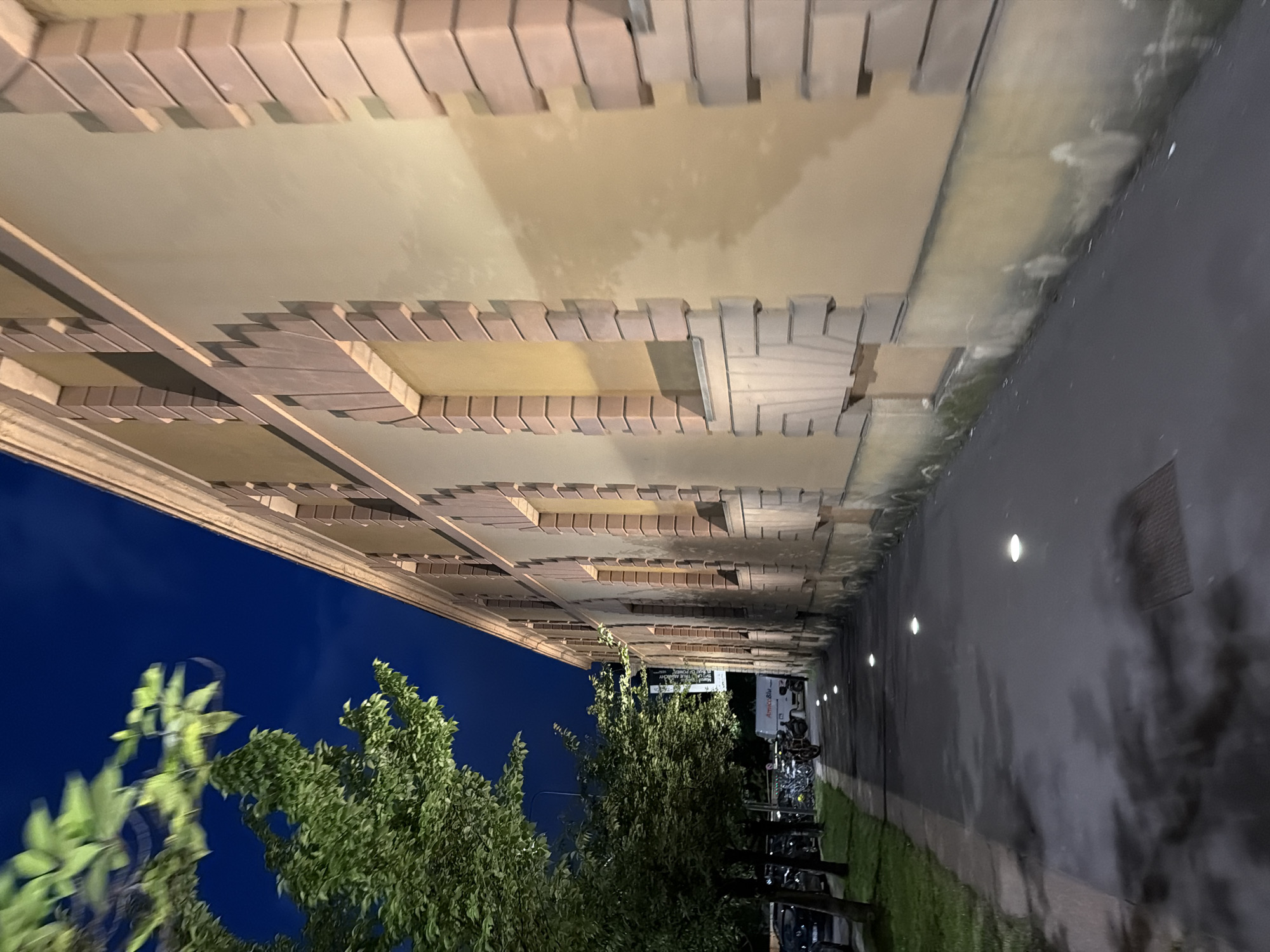 This image shows a building exterior in Milan, Italy. The building has a brick facade with a patterned design. A dark blue sky can be seen through the trees in front of the building. There are trees to the left and in the mid-ground. A sidewalk runs along the building, with a drain. The sidewalk is lit by ground-level lights. There are parked motorcycles, scooters, and cars in the background. People are present near the end of the buildings.