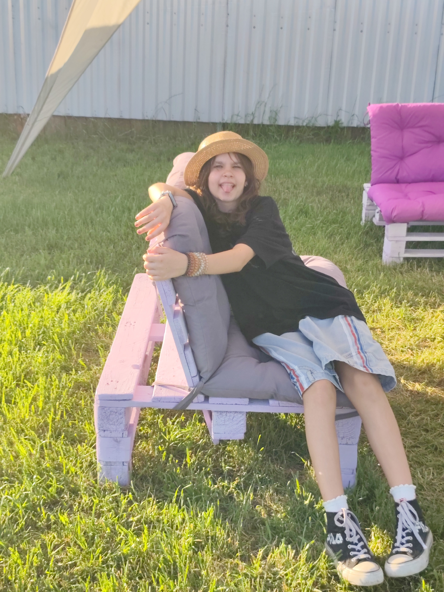 A young person is lounging on a pink-painted pallet chair outdoors in the grass. They are wearing a straw hat, a black t-shirt, light blue shorts with red and white stripes, and black high-top sneakers with white laces and white ruffled socks. The person is sticking their tongue out playfully at the camera, with their arms resting on the chair and bracelets on one wrist.
In the background, there is a white corrugated metal fence, a partially visible light-colored tarp or tent, and another pink pallet chair with a cushion. The scene is set in a grassy area, likely a yard, with the sun casting long shadows, suggesting it is late afternoon or early evening. The lighting is warm and the grass appears green and lush, indicating a pleasant weather day. The location is Nechaiane, Ukraine. There is no visible text in the image.