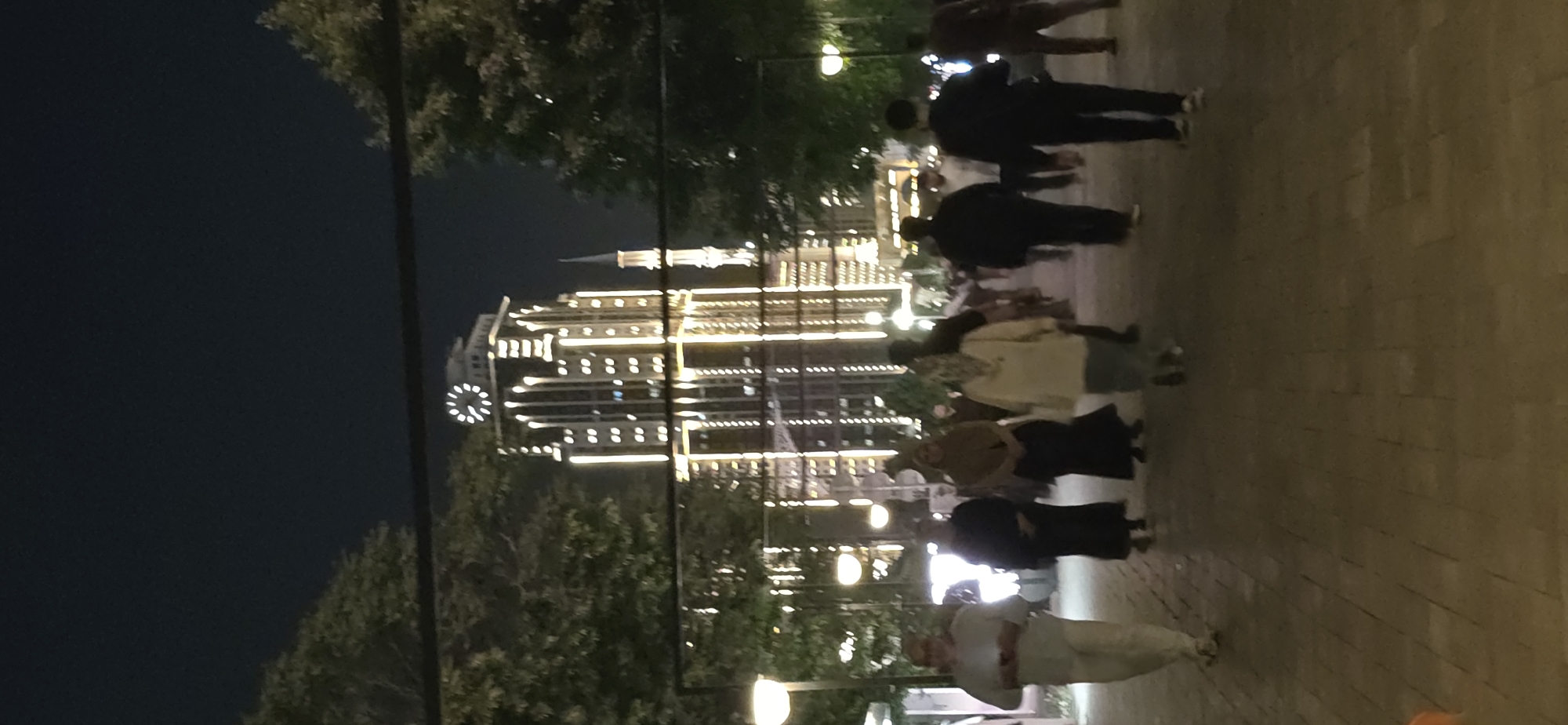 At night in Grozny, Russia, a lit building stands amidst trees. Several people walk on a paved walkway. The image is blurry and dark.
