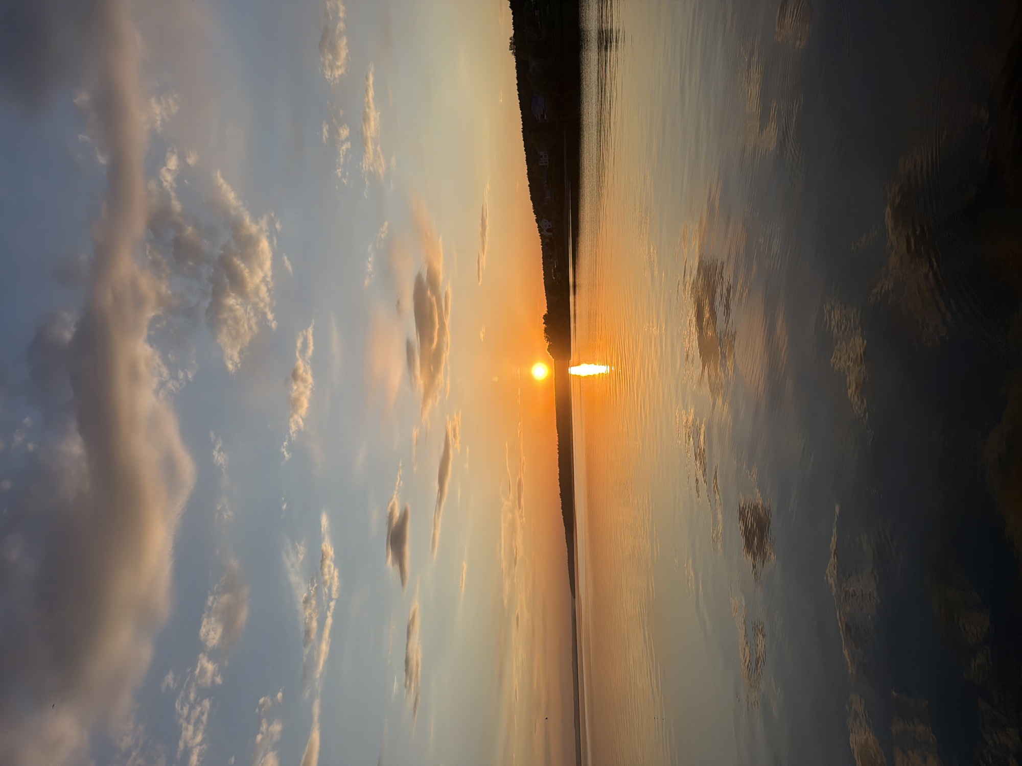 The image depicts a sunset over a body of water, likely a lake or wide river. The sun is positioned just above the horizon line, casting a bright, orange glow across the water and sky. A thin strip of land, possibly a shoreline with trees, is visible in the center of the image. The sky contains scattered clouds, some reflecting the colors of the setting sun. The water's surface reflects the sky and sun, creating a mirrored effect. Given the prompt, it is plausible that this location is near Votkinsk, Russia. No people are visible in the image.