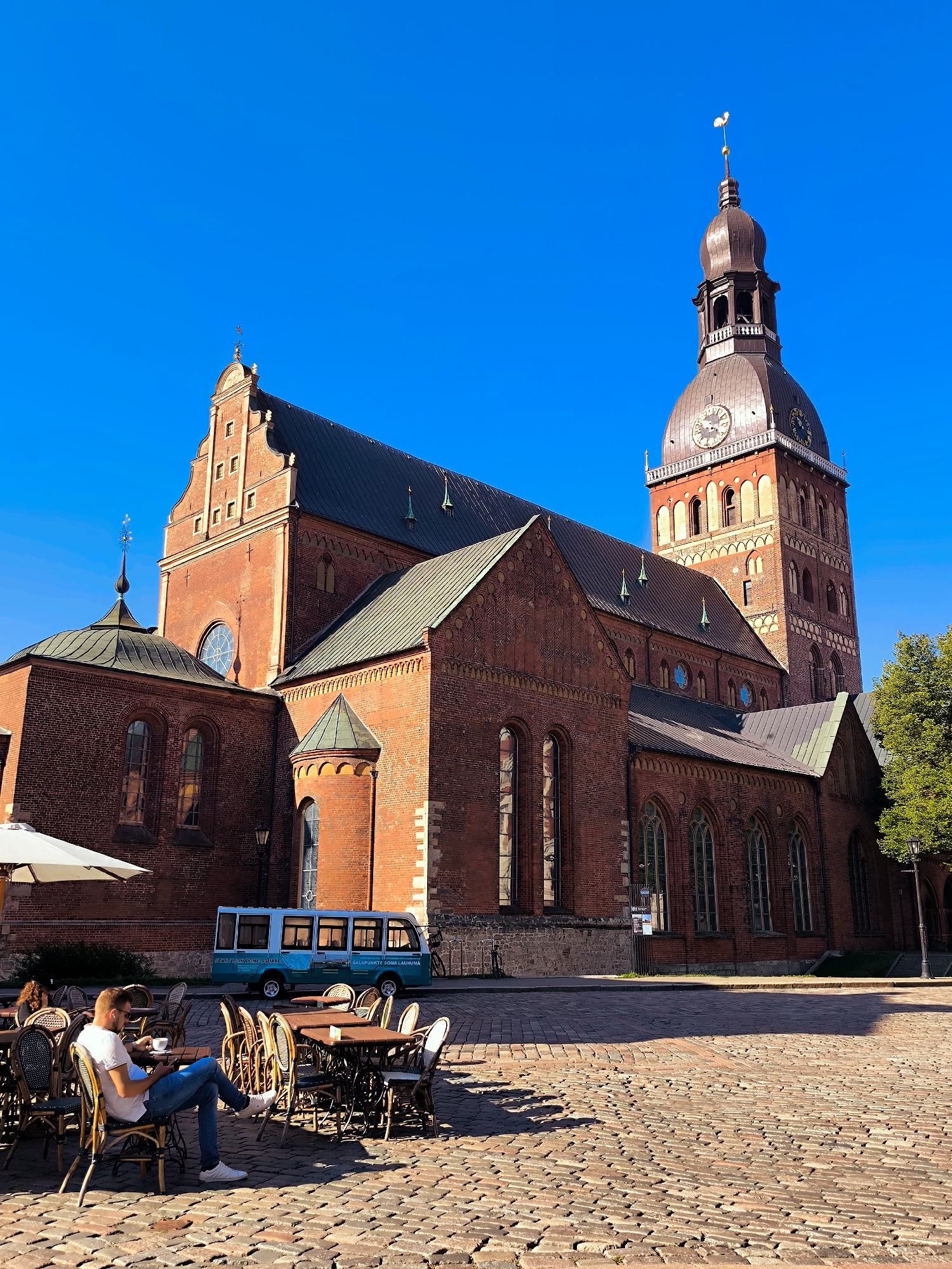 The image depicts a sunlit scene in Riga, Latvia, specifically in the Doma Square (Daugavas). In the background stands the Riga Cathedral, a prominent brick church with a tall, dark tower featuring a clock face. The cathedral's architecture is characterized by its red brick facade, arched windows, and pitched roofs. In the foreground, a cobblestone plaza is laid out with numerous outdoor tables and chairs belonging to a cafe. A man, casually dressed in a white t-shirt and blue jeans, sits at one of the tables, engrossed in his mobile phone. He has a cup of coffee on the table. Next to him, another table is occupied by a partially visible person. A light blue tourist bus with "DOME SQUARE" written on its side is parked on the plaza behind the tables. An umbrella provides shade over some of the seating. The sky is a clear, vibrant blue, and the overall impression is one of a pleasant day in a historic urban setting.