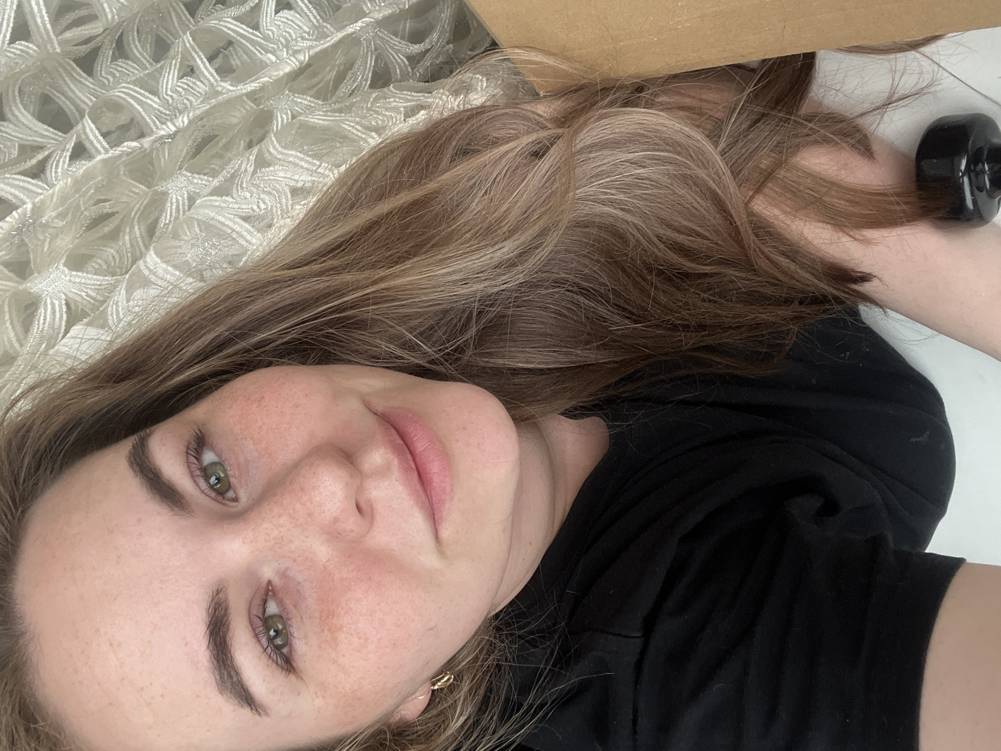 A woman with freckles lies on her side, partially under a cardboard box. She has fair skin, light brown hair with highlights, and green eyes. She wears a black shirt. Her head rests on a white, lacy fabric. There is a black object near her arm. The location and city of Chaikovsky, Russia are unknown.