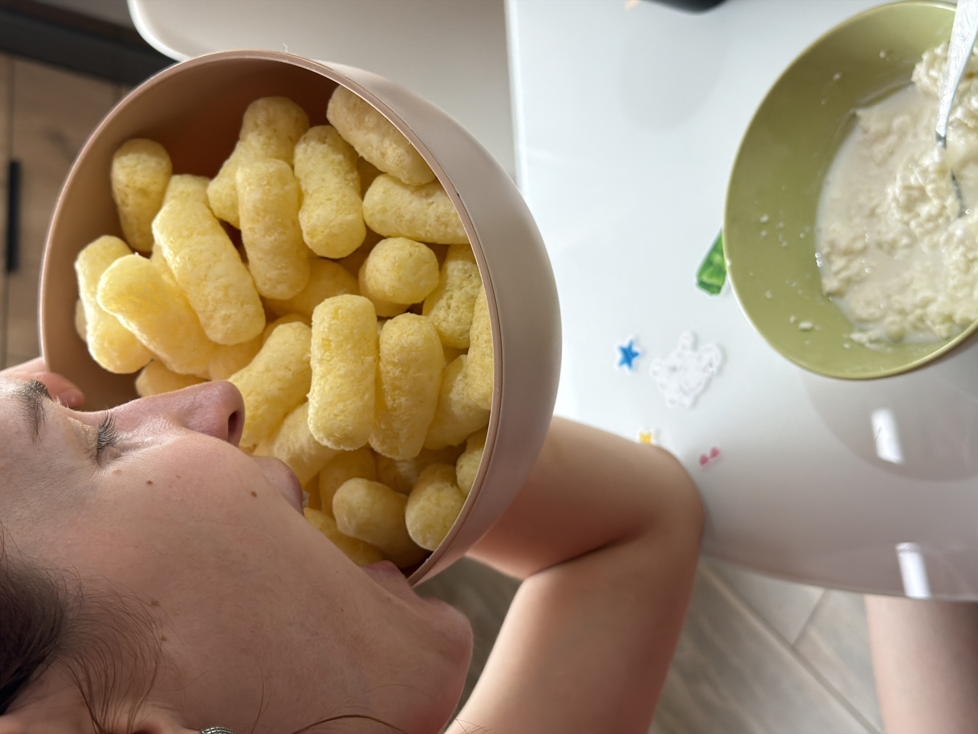 A close-up image captures a person's profile, from the nose downward, consuming yellow corn puffs directly from a rose-toned bowl. The individual has light skin and visible eyelashes. A portion of their left arm is visible as they hold the bowl. The bowl is positioned over a white surface, identified as a table. On this table, to the lower right, a light green bowl contains a white, lumpy substance consistent with cottage cheese submerged in milk, with a metal spoon resting within it. Small decorative elements, including a blue star and white cutouts, are affixed to the white table surface. The flooring visible in the lower left exhibits light-colored, plank-style tiling. The background is out of focus, indicating an indoor environment. No visual information within the image confirms the specified location as Chaikovsky, Russia.