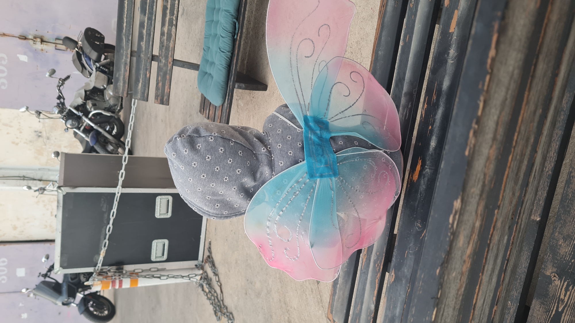 A person with their back turned and wearing a gray patterned hat sits on a wooden bench, with pink and blue fairy wings attached to their back. The bench appears to be located outdoors. A motorcycle and a road case secured with a chain are visible in the background. The location is Rishon LeZion, Israel, as indicated by the "306" marking on the case.