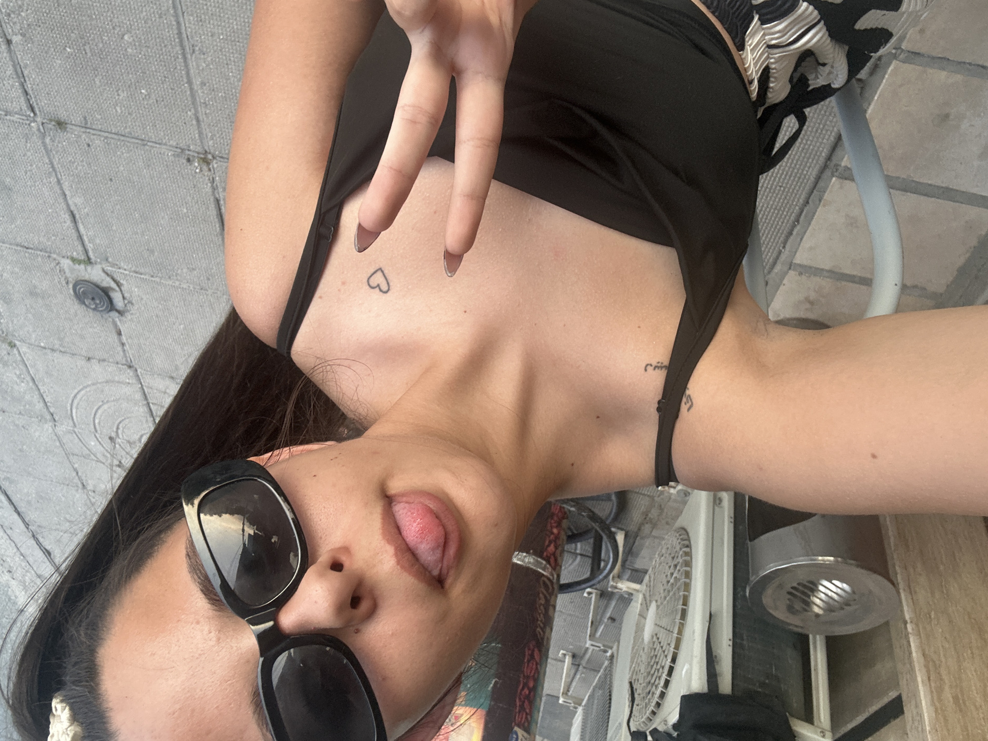 A young woman with dark hair and a light to medium skin tone captures a selfie, holding her right hand up in a peace sign with manicured nails. She wears black sunglasses, a black spaghetti strap top, and patterned black and white bottoms. Her tongue is slightly extended, and she has a small outline of a heart tattooed on her upper chest, along with Arabic script on her left shoulder/upper arm. A light-colored, textured hair accessory is visible in her hair on the right side of her head. The background indicates an outdoor setting with grey rectangular paved ground. To the left, a tall, silver cylindrical ashtray or public bin stands next to a wooden panel. An outdoor air conditioning unit with a visible fan grille is situated behind her on the right. Parts of a light-colored metal chair are also visible in the mid-ground. The reflections in her sunglasses show an outdoor scene, possibly with sky and structures. The location is Unknown, Unknown.