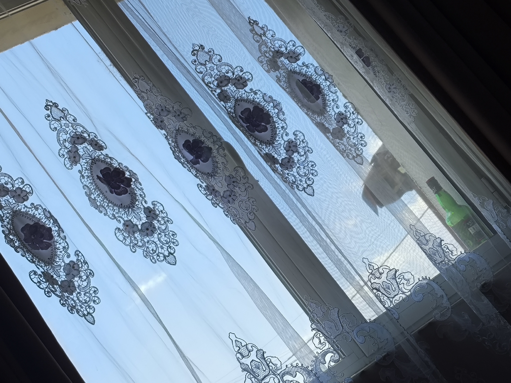 This image depicts an interior scene in Boliartsi, Bulgaria, viewed through a window partially covered by sheer curtains. The primary subjects are several translucent white or light-colored curtain panels, adorned with repeating embroidered patterns. These patterns consist of dark purple or blue intricate lacework, featuring large oval medallions centrally housing a darker, four-petal floral motif, flanked by smaller floral and scroll designs.
Behind the curtains, a window frame is discernible, suggesting a multi-pane or multi-section window. Beyond the window, a clear, bright blue sky is visible, indicating a daytime setting. On the right side of the image, near the top of the window sill, a light-colored, indistinct silhouette of an animal, possibly a cat, is faintly visible through the sheer fabric. Further to the right, on the window sill, a green glass bottle with a dark label is partially obscured by the curtain. Darker, heavier curtains or blinds are visible along the far left and right edges of the frame, partially framing the sheer curtains and the window. The scene is illuminated by natural light entering from the exterior.