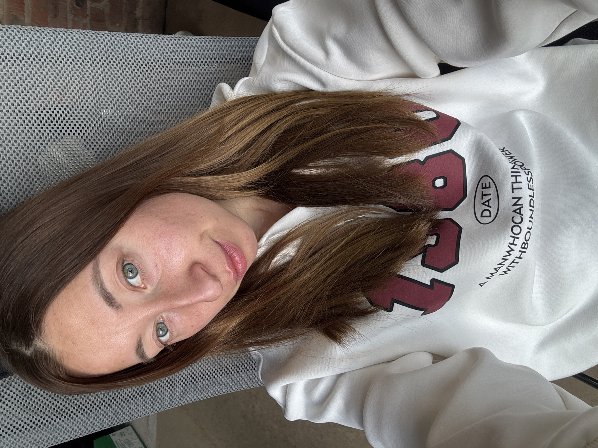 A fair-skinned woman with light brown hair and grey-blue eyes is lying on a grey mesh chair. She is wearing a white hooded sweatshirt with a maroon number and the word "DATE" printed on it. The sweatshirt also has the words "A MAN WHO CAN THINK WITH BOUNDLESS POWER" printed on it. The background includes a brick wall. The location is not discernible as Chaikovsky, Russia.
