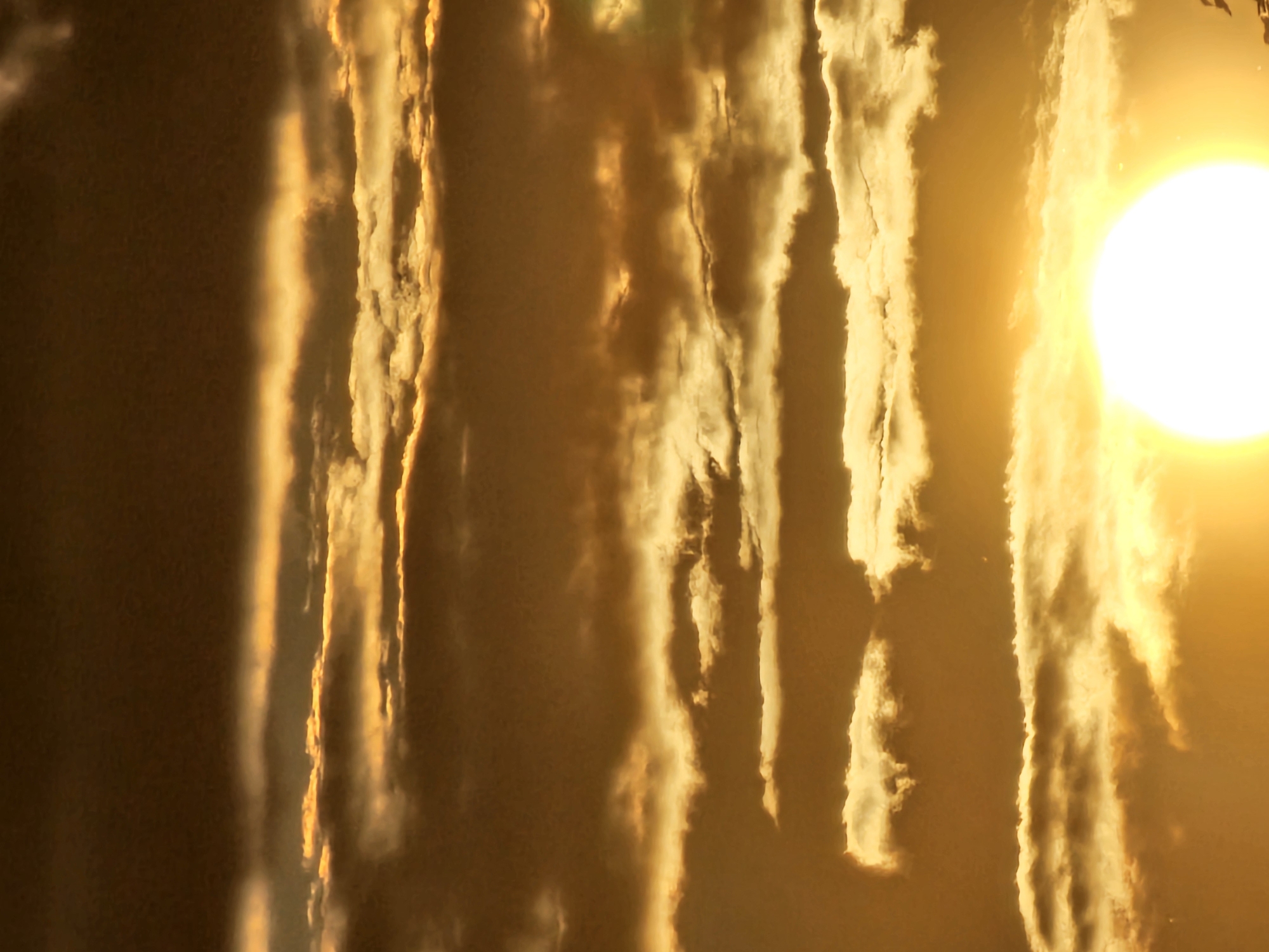The image is a close-up, vertical shot of a dramatic sunset over Khimki, Russia. The sky is filled with layers of clouds, illuminated by the bright, white sun located in the bottom right quadrant of the frame. The clouds range in color from deep brown at the top to vibrant gold and orange closer to the sun. The sunlight is intense, creating a radiant glow that permeates the entire scene. There are no visible people, subjects, objects, or actions, only the natural environment of the sky and clouds during sunset.