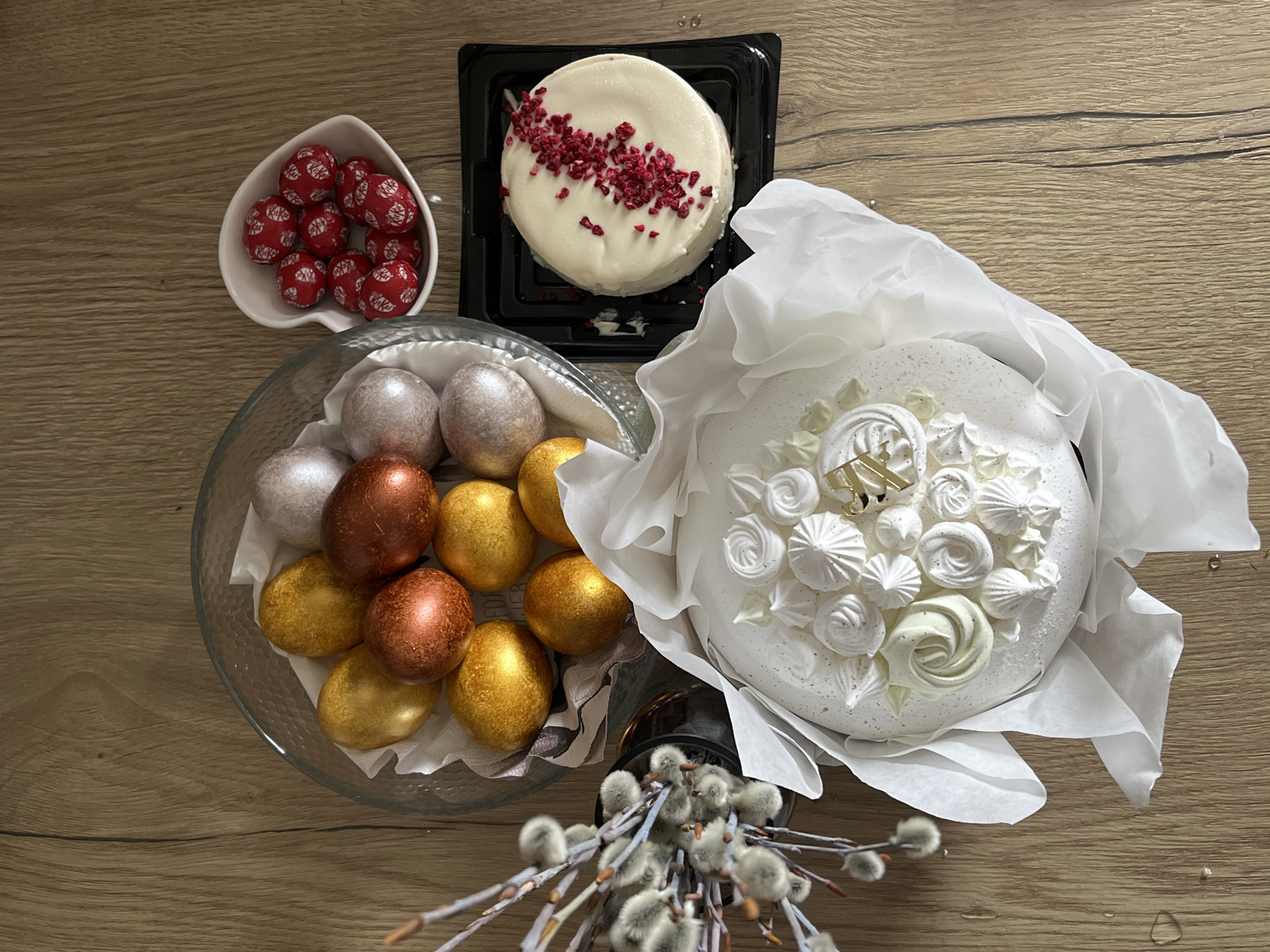 The image shows a collection of Easter treats arranged on a light brown wooden surface. A heart-shaped white bowl contains red KitKat chocolate balls. Next to it, a glass bowl holds Easter eggs in metallic shades of silver, gold, and bronze. A small white cake topped with a white glaze and sprinkled with red dried raspberries sits on a black square plastic container. Another larger white cake, decorated with white meringue swirls and a gold cake topper, is partially wrapped in white tissue paper. A small bouquet of pussy willow branches is at the bottom of the image. The location is not provided by the image.
