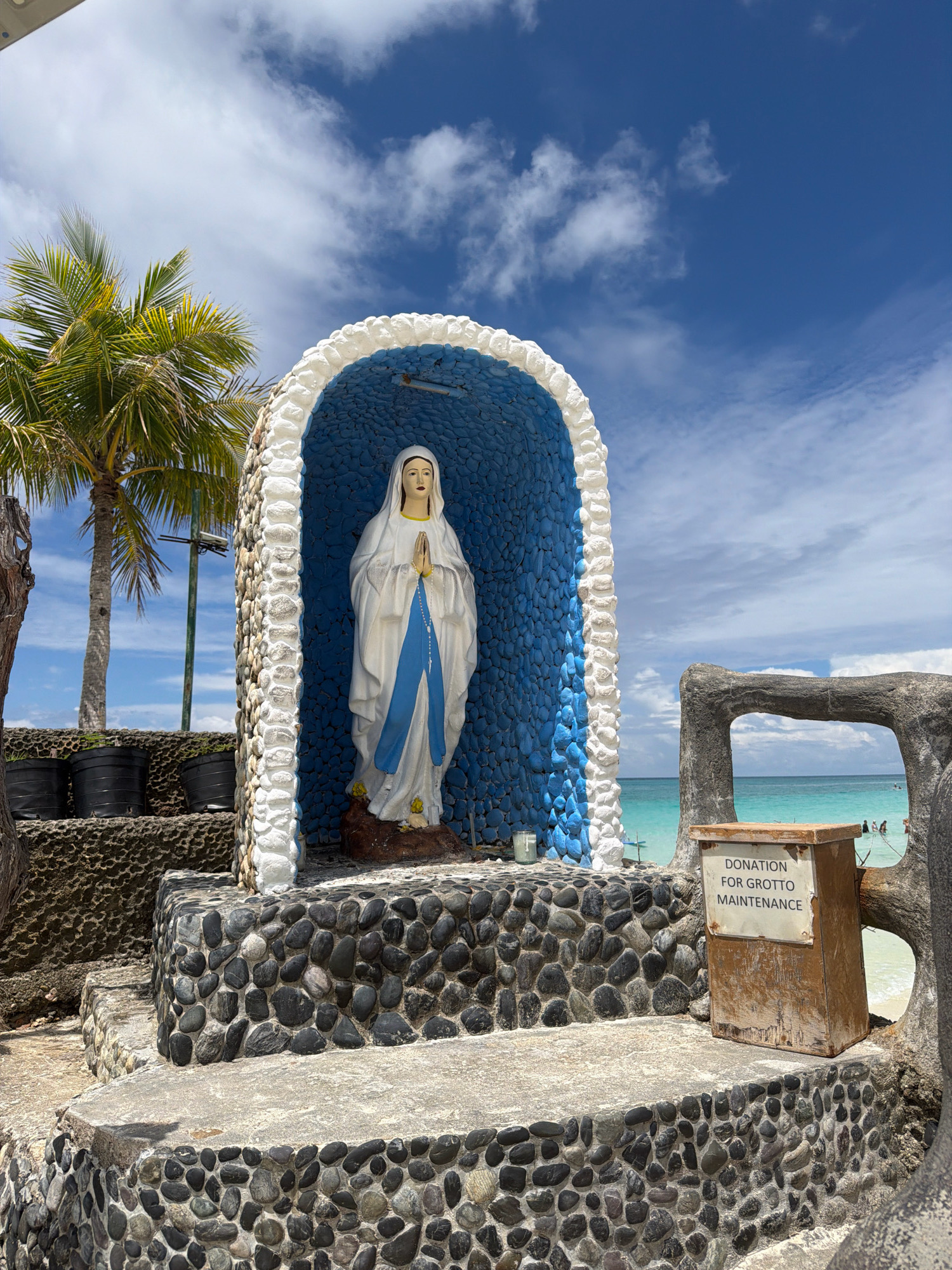 A statue of the Virgin Mary stands within a grotto decorated with blue tiles and a white stone arch, located outdoors on a sunny day in Malay, Philippines. The grotto is situated on a multi-tiered stone structure, with a donation box labeled "DONATION FOR GROTTO MAINTENANCE" placed to the right of the steps. Behind the grotto, a clear turquoise sea is visible with a few people in the water. A palm tree stands to the left of the grotto, and the sky is filled with white clouds.