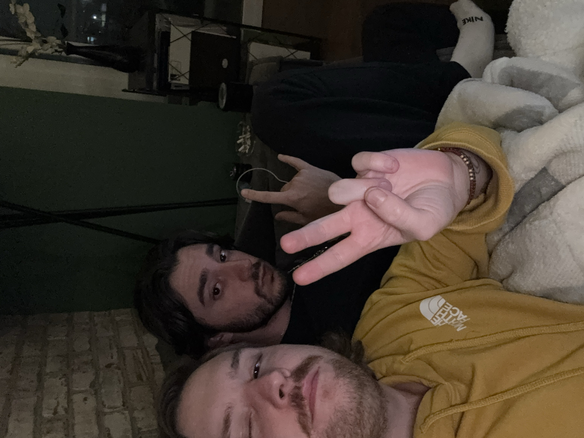 Two men are laying down, with the man closest to the camera wearing a gold colored North Face sweatshirt and the man behind him wearing black. Both men have beards. A white Nike sock is visible in the background. Both men are making hand gestures. It is not possible to determine the location.
