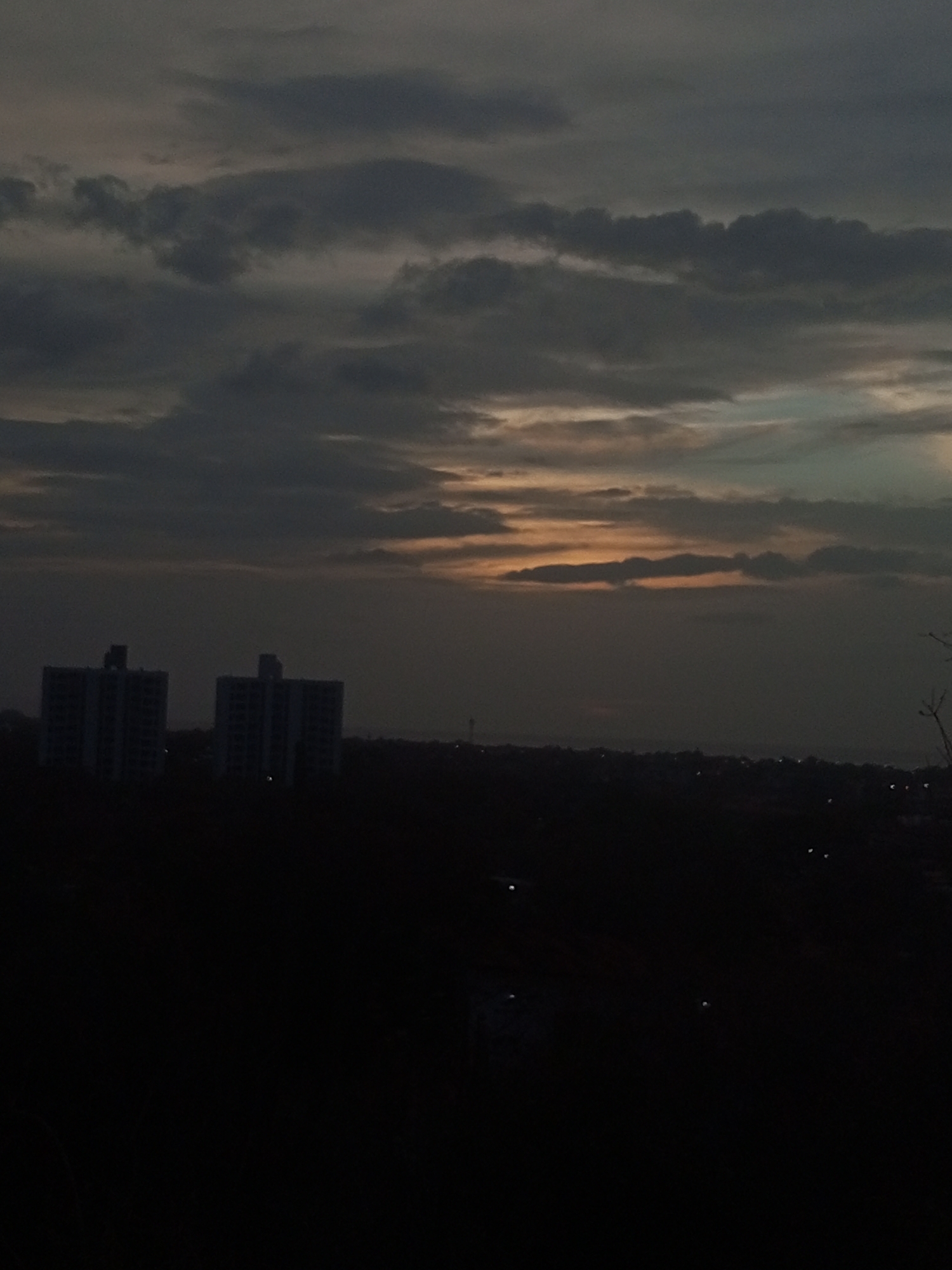 The image shows a dark, overcast scene in Altagracia, Venezuela. In the distance, two tall buildings are silhouetted against the dim light of the horizon. Below them, a dark, undifferentiated area suggests a forest or dense vegetation. Above, heavy clouds dominate the sky, with a hint of orange light breaking through near the horizon. There are no visible people or interactions in the scene. The overall environment is gloomy and suggests either late evening or early morning.