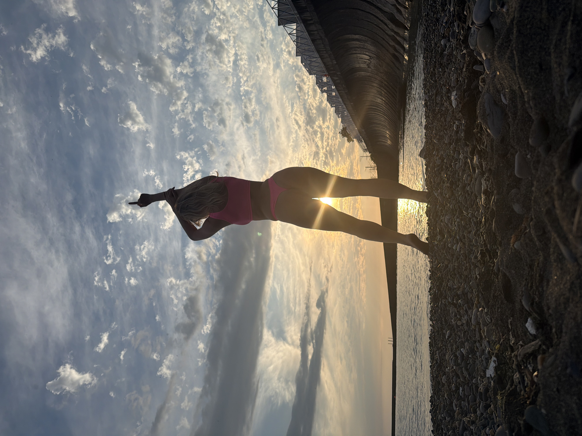 A person in a pink bikini stands on a pebble beach next to the sea, with a sunset creating a dramatic silhouette. The person is facing away from the camera, with their arms extended and one hand raised, as if pointing. The sun is positioned behind the person, creating a lens flare that obscures their face and highlights their form. The sky is filled with fluffy white clouds, catching the golden light of the setting sun. To the right of the person, a dark, curved structure stretches into the distance, possibly a pier or a man-made barrier along the coastline. The water is calm and reflects the warm tones of the sky. The overall mood is serene and possibly empowering, with the natural beauty of the sunset enhancing the solitary figure. The image appears to be taken in Sochi, Russia, given the context provided.