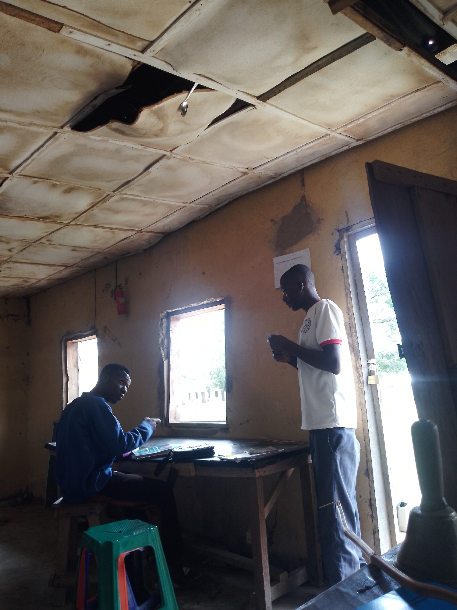The image depicts the interior of a sparsely furnished room, likely a classroom or workshop, with two young men present. The ceiling is in disrepair, with large sections missing, revealing the darkness of the space above. One man, wearing a blue long-sleeved shirt and dark pants, is seated at a wooden desk, looking down at papers or books. A colorful plastic stool is positioned in front of him. The second man, wearing a white t-shirt with red trim and dark pants, stands near a doorway, holding a small object in his hands and looking to his right. A fire extinguisher is mounted on the wall to the left of a window, and another window is visible on the far right, with a partially open door beside it. The walls appear to be made of earth or mud plaster, and the overall condition of the room suggests neglect or a lack of resources.