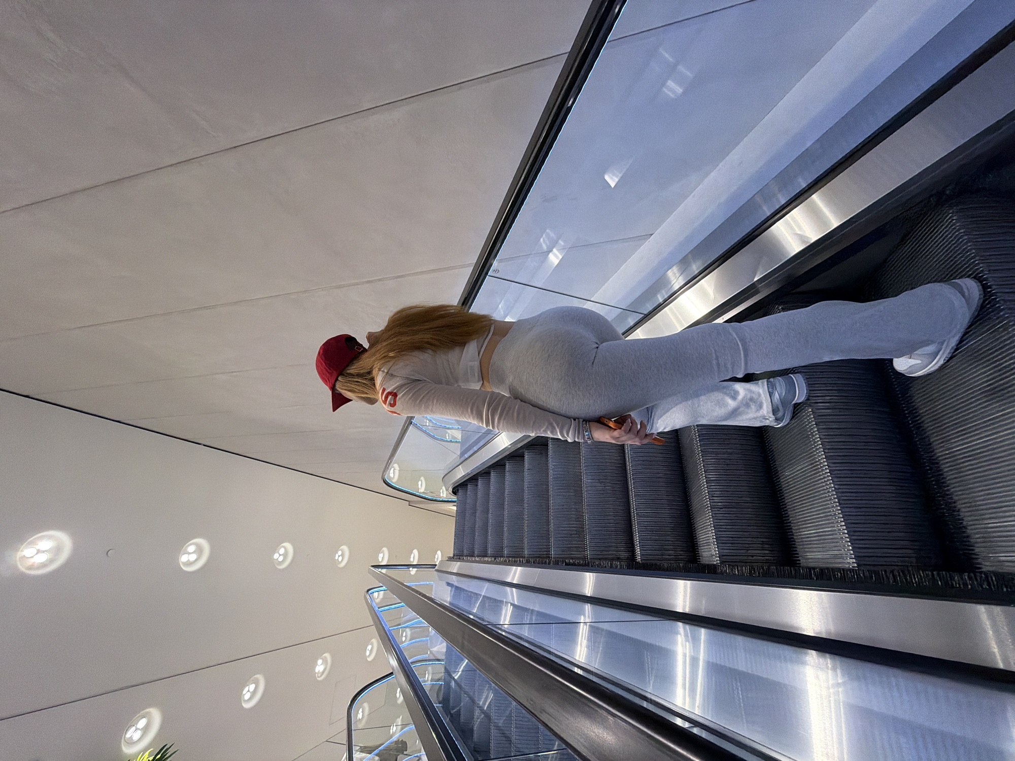 A woman with blonde hair and a red baseball cap is lying face down on an escalator. She is wearing a white long-sleeved shirt and gray leggings. She is holding a cell phone in her hand. The escalator is located in a modern building with a white wall and circular lights. The building is possibly located in Sofia, Bulgaria.