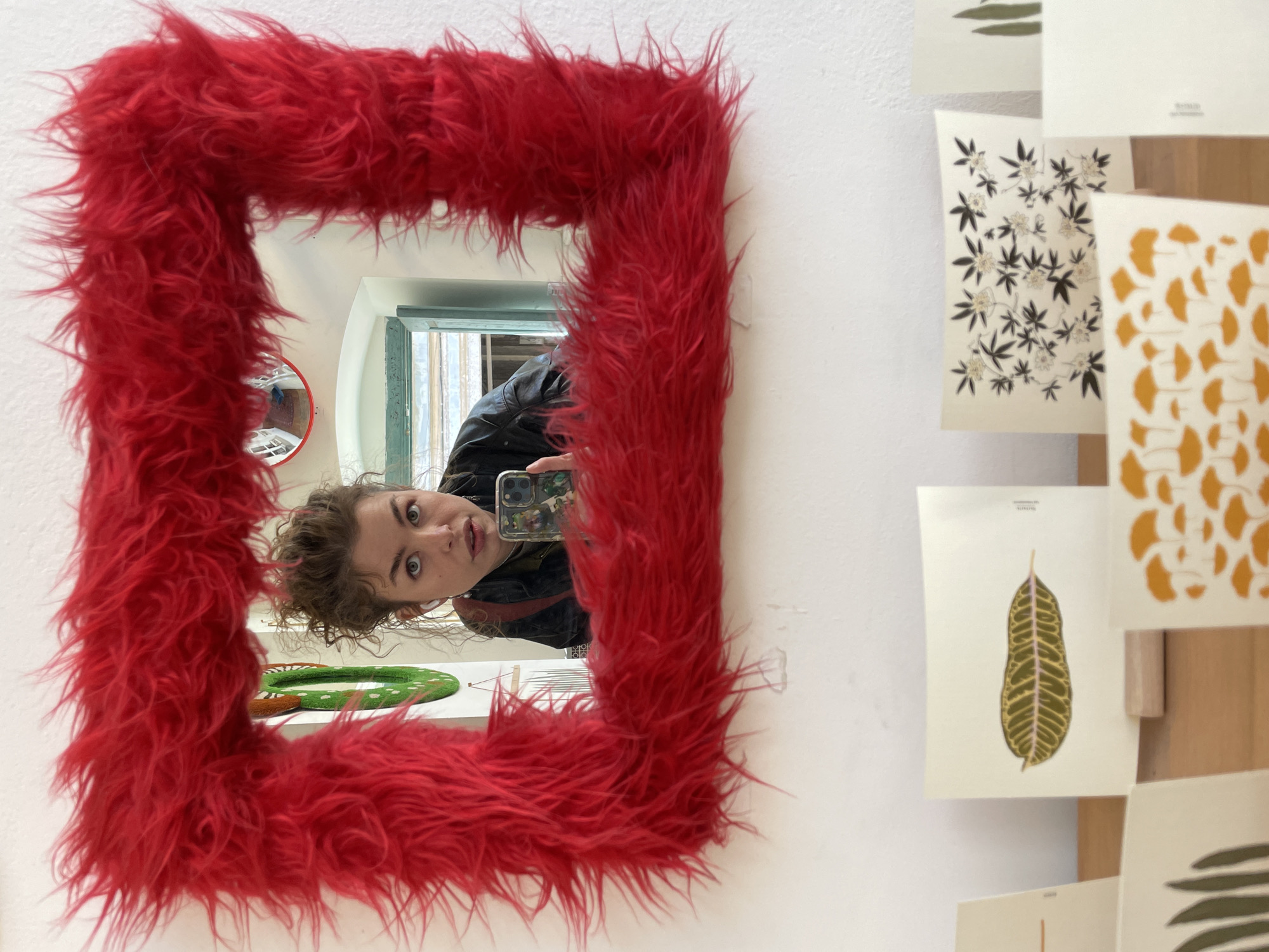 A woman takes a selfie in a mirror with a red, furry frame in Tbilisi, Georgia. The mirror reflects the woman holding a phone, as well as a glimpse of a room with various objects. Adjacent to the mirror is a display of botanical print cards.