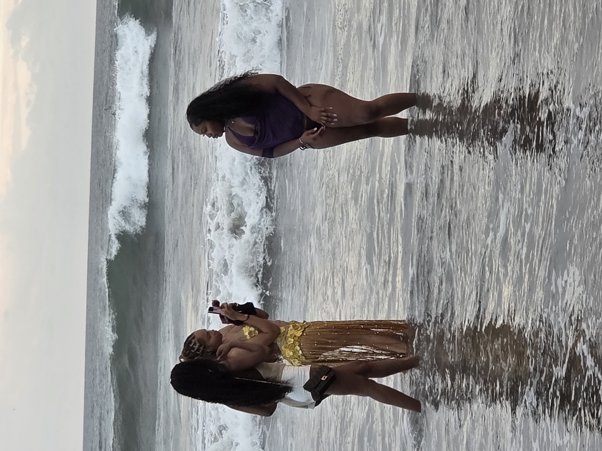The image captures two individuals on a beach in North Kuta, Indonesia, likely during the late afternoon or early evening given the soft, diffused light. The scene is dominated by the ocean, with gentle waves lapping at the shore and reflecting the sky.
The first individual, positioned closer to the foreground and to the right of the frame, is wearing a dark, one-piece swimsuit. They have long, dark hair and appear to be looking down, possibly at their phone, which they are holding in their hand. Their pose suggests a relaxed or contemplative mood.
The second individual, situated further back and to the left, is wearing a shimmering gold garment, possibly a skirt or dress, paired with a white bottom and a dark handbag. This person is also holding a phone and seems to be interacting with the first individual, or perhaps posing for a photo. Their hair is styled in braids with some loose strands.
The water is shallow, covering the legs of both individuals, and the wet sand reflects the shimmering gold of the garment. The overall atmosphere is serene and peaceful, with the natural beauty of the beach providing a tranquil backdrop. There is no visible text in the image.