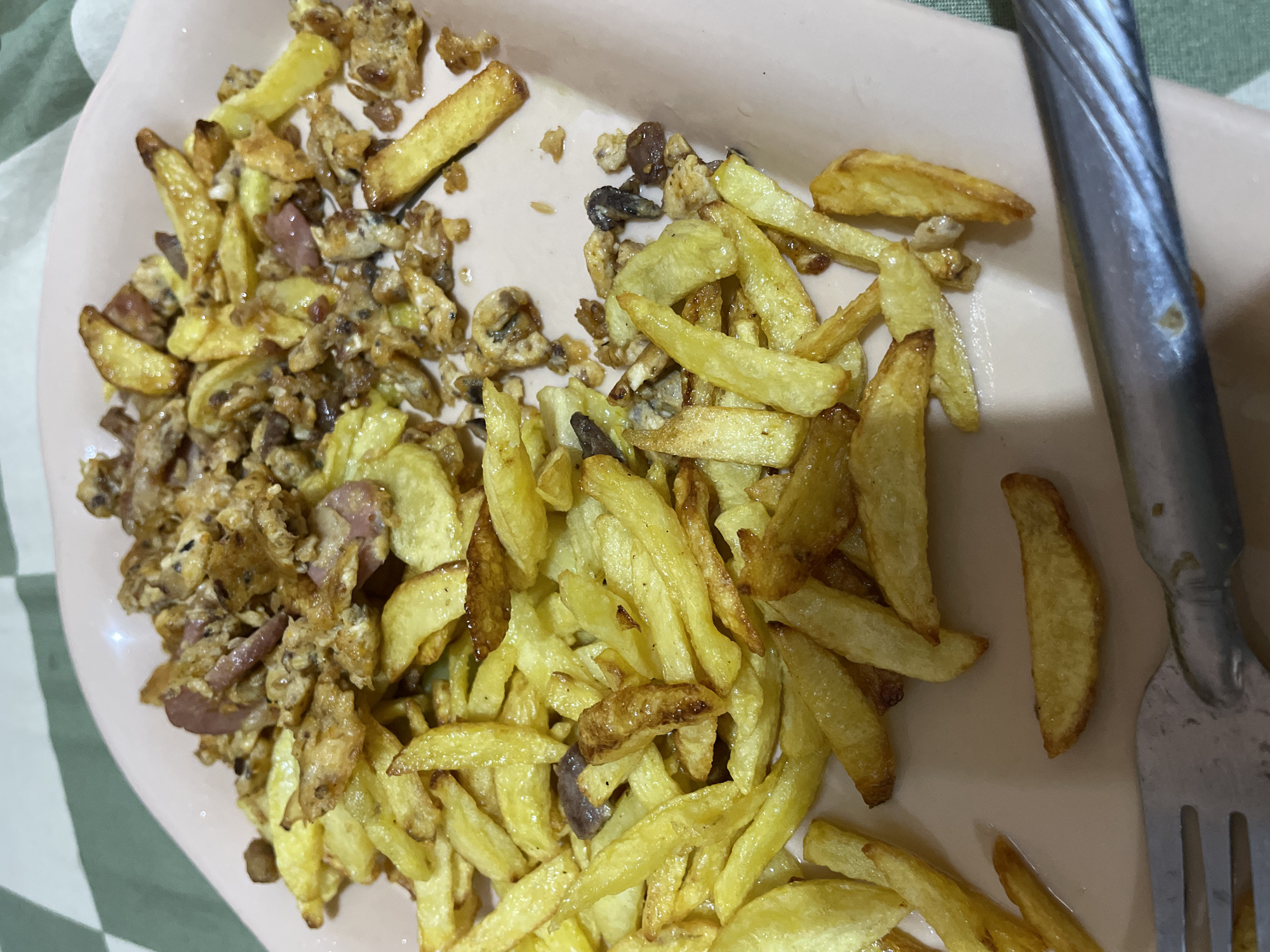 This image features a close-up shot of a plate of food, likely taken indoors. The plate is a pale pink, oval dish containing a generous serving of French fries mixed with what appears to be scrambled eggs and possibly pieces of sausage or other meat. The food is golden brown and looks freshly prepared, with a slightly greasy sheen.
The French fries are cut into thick, steak-fry style pieces and are scattered throughout the dish. Interspersed with the fries are crumbles of scrambled egg, some small, dark brown pieces that could be spices or herbs, and reddish-pink slivers that suggest bits of meat.
To the right of the plate, a portion of a silver fork is visible. The fork's tines are slightly blurred, indicating it's close to the edge of the frame and possibly being used to eat the meal. The background is partially visible, showing a green and white checkered tablecloth or mat, suggesting a casual dining setting.
There is no visible text in the image. The lighting appears to be artificial, consistent with an indoor meal. The overall impression is of a hearty, savory meal.