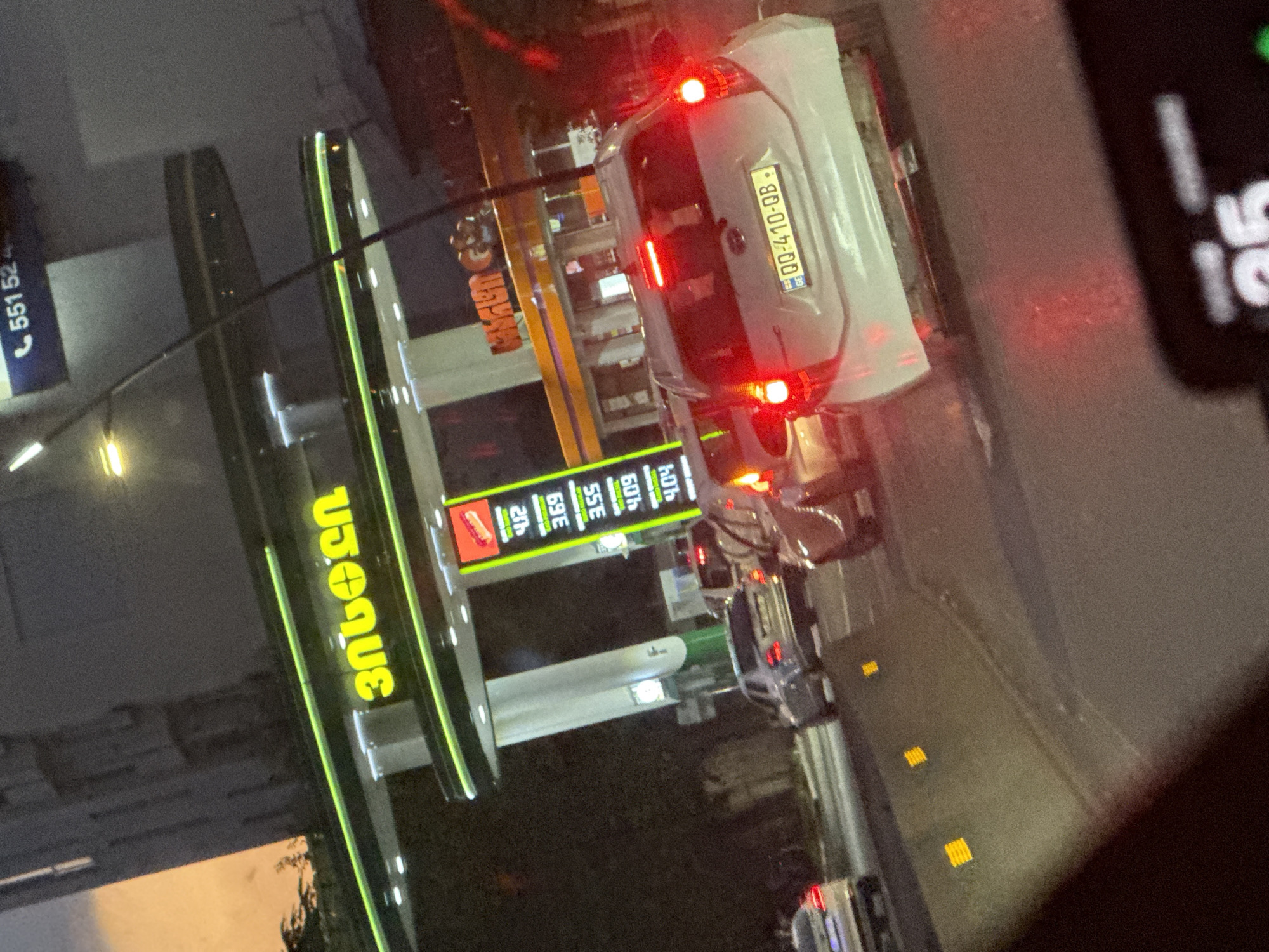 The image depicts a traffic scene at night in Tbilisi, Georgia, specifically at a Rompetrol gas station. The gas station features bright green and yellow signage with the Rompetrol logo and Georgian lettering that reads "რომპეტროლი" (Rompetrol). A digital display at the gas station shows fuel prices: 4.12, 3.69, 3.55, 4.09, and 4.04. Several cars are visible in traffic, including a white Toyota Yaris with license plate QQ-410-QB, a silver Mercedes-Benz C-Class, and other vehicles partially obscured. The sky is a dusky orange, suggesting either dawn or dusk. The scene is lit by the gas station's illumination and the headlights and taillights of the vehicles.