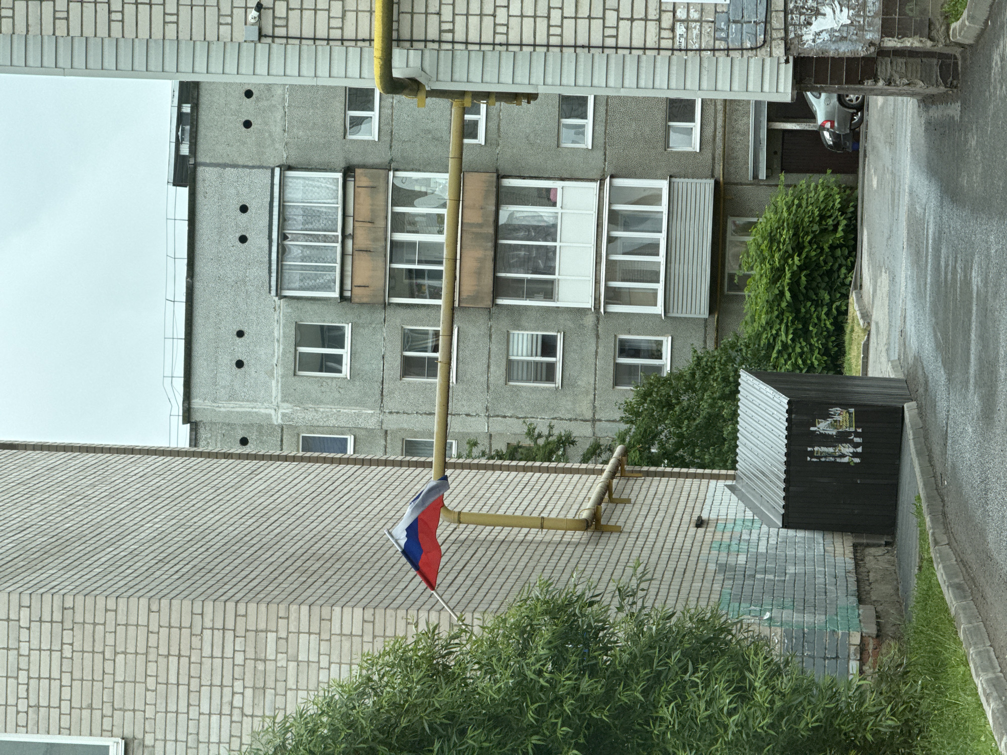 This image depicts an exterior view of an apartment building and a small, dark structure with a metal roof. A Russian flag is visible, attached to a pole and waving gently. The flag is positioned above a dense green shrubbery and a sloped, tiled roof of a building. A yellow pipe runs diagonally across the scene, attached to the apartment building.
The apartment building is made of grey concrete and has multiple floors with numerous windows. Some of the windows are covered with blinds or curtains. A white balcony with glass panels is also visible. In the background, a silver car is parked near an entrance to the building. The ground is paved with asphalt and a grey curb is present. Green grass and trees are scattered throughout the scene.
The image appears to be taken during the daytime, under an overcast sky. The lighting is diffused, suggesting a cloudy or possibly rainy day. The overall scene conveys a sense of an everyday urban or suburban environment. No people are visible in the image. No text is discernable in the image.