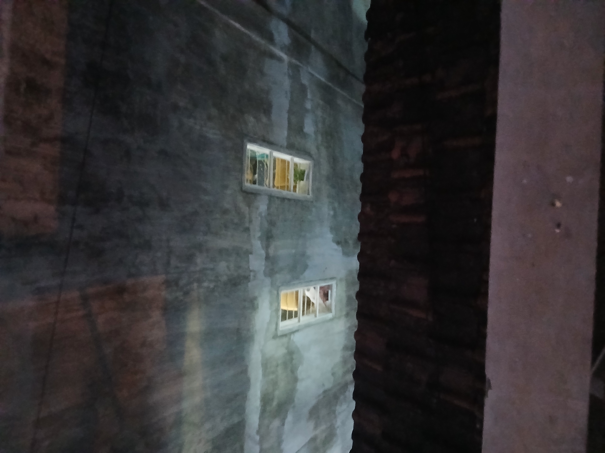 The image shows an exterior view of a building's concrete wall with two small rectangular windows. The windows are partially illuminated from within, with one window appearing to have some fabric or bedding visible. A dark, corrugated roof structure is in the foreground, suggesting the viewpoint is from below or an adjacent lower structure. The overall scene is dimly lit, hinting at dusk or nighttime. The location is identified as Tenggarang, Indonesia, though no specific landmarks or contextual details within Tenggarang are discernible from the image itself.