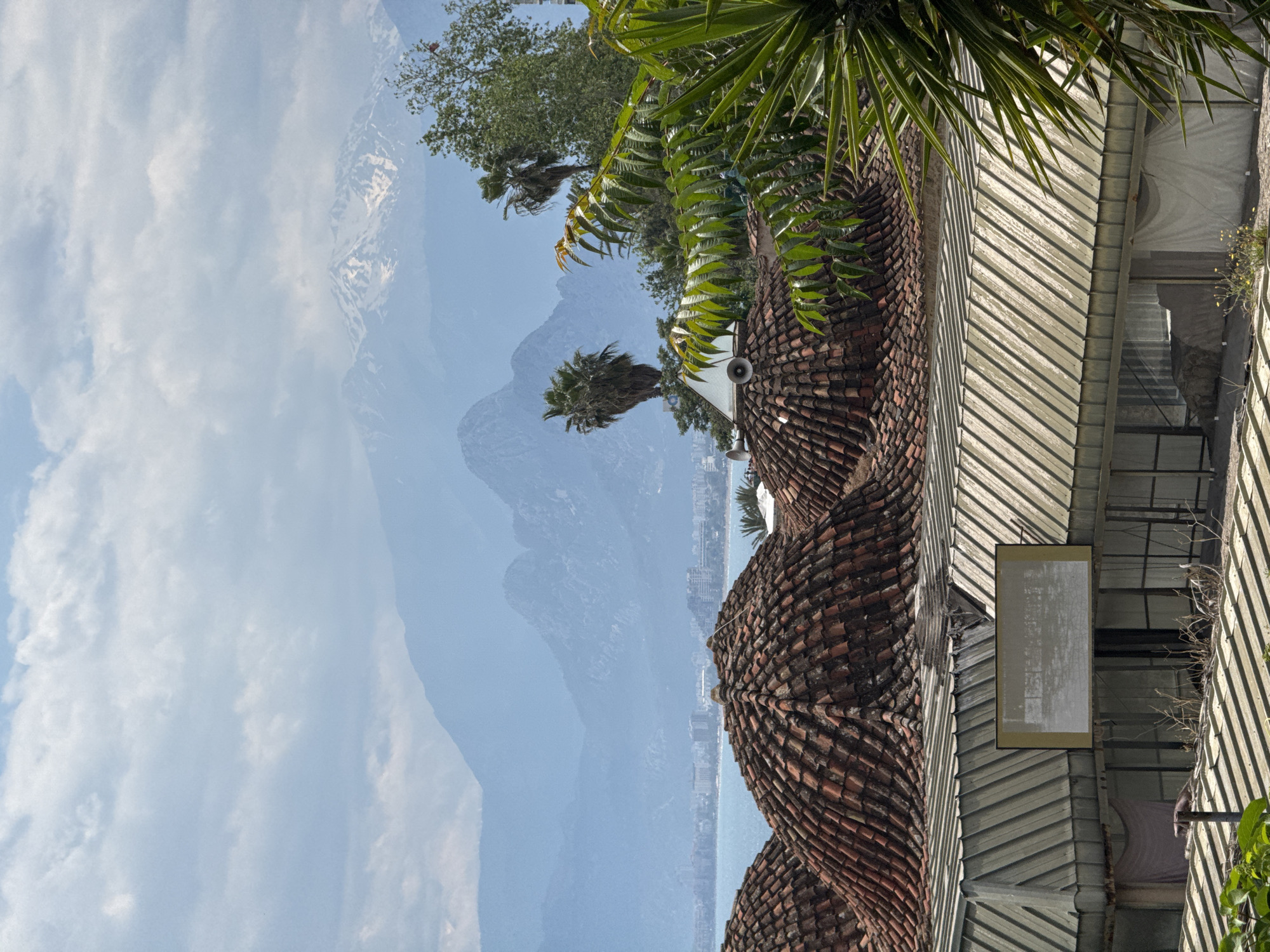 The image shows a view of mountains and the city of Antalya, Turkey, seen through foliage and over tiled domes. The landscape includes mountains in the background with some snow-capped peaks, partly obscured by clouds. The city of Antalya appears as a line of buildings along the coast.
The foreground includes the tiled domes of a building, possibly a mosque or other traditional structure. A palm tree and other foliage are visible, partially framing the mountain view. A metal roof and sign are also present.