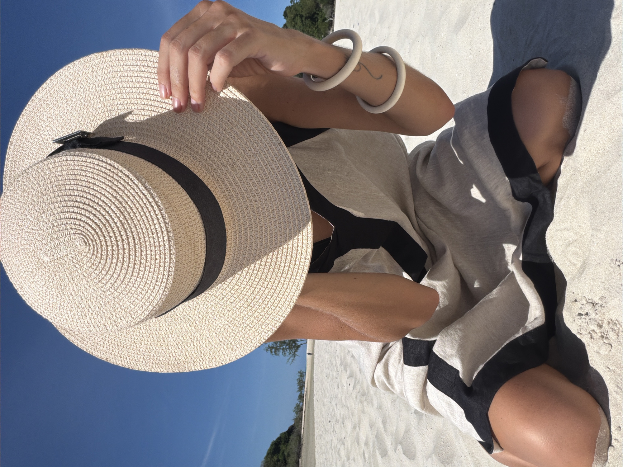 A person sits on a sandy beach under a clear blue sky, wearing a wide-brimmed straw hat that obscures their face. They are dressed in a loose, light-colored garment with black trim. The person's hands are visible, with one hand holding the hat and the other resting on their arm, adorned with two light-colored bangles. The background shows a shallow body of water, vegetation on a distant shore, and a single figure walking in the distance. The sand is light-colored and appears fine-grained. The scene is brightly lit by sunlight. The location is specified as Baan Plai Laem, Thailand.
