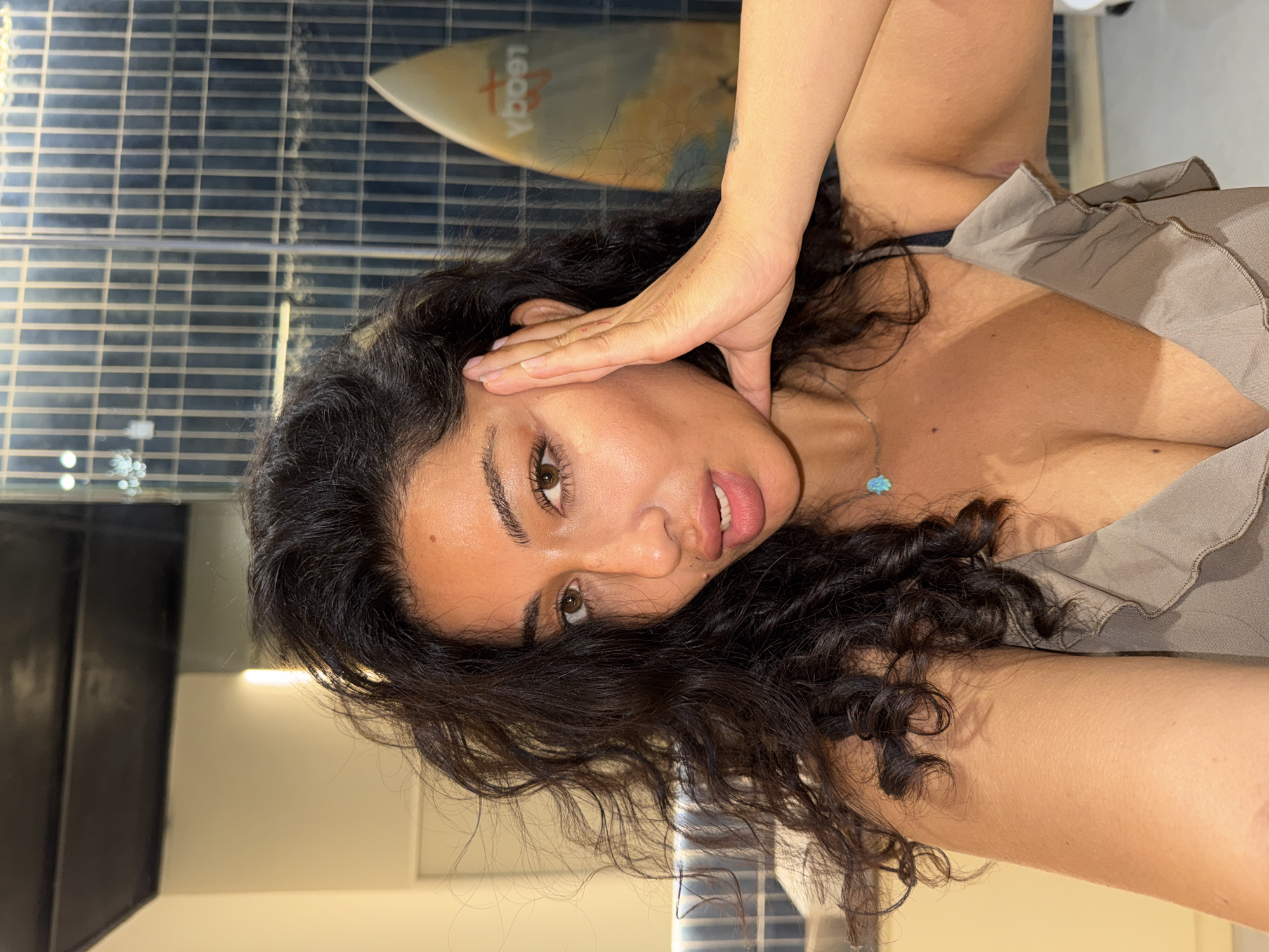 The image displays a head-and-shoulders portrait of a young woman, captured from a slightly high angle and rotated counter-clockwise. She has long, dark, curly hair and light brown eyes, looking directly towards the viewer with a relaxed expression and slightly parted lips. Her left hand is raised, with fingers gently splayed, resting against her temple and partially obscuring her forehead.
She is wearing a light brown or taupe-colored ruffled top, revealing her décolletage, and a visible black bra strap on her left shoulder. A delicate silver necklace with a small turquoise pendant adorns her neck.
The background features a tiled or patterned wall with horizontal and vertical lines, giving a grid-like appearance. Partially visible in the upper left (when upright) is a light-colored surfboard with the reversed text "YOOBAA" printed on it. Reflections of lights or windows are visible in the background, suggesting an indoor or semi-enclosed setting. The presence of the surfboard, along with the given location context of Kuta Selatan, Indonesia, hints at a coastal or surf-related environment, possibly a hotel, cafe, or private residence. The lighting appears bright and even, indicating daytime or a well-lit interior.