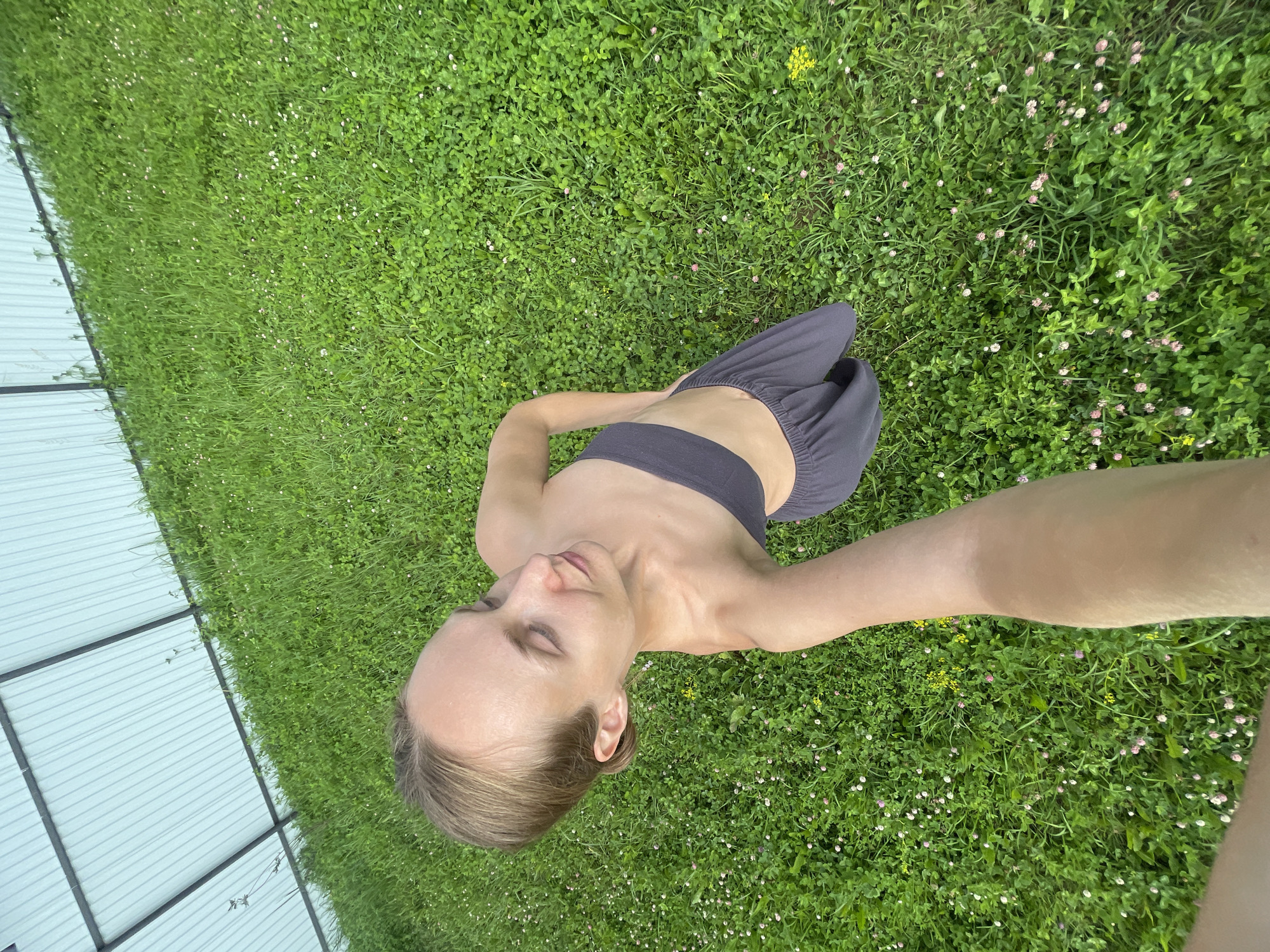 This media file is a high-angle, wide shot of a person lying on a grassy field with a corrugated metal structure to the left. The person, a fair-skinned individual with blonde hair pulled back, is wearing a dark, strapless top and dark pants. Their eyes are closed, and their head is tilted back, suggesting a state of relaxation or meditation. The grass is lush and green, dotted with small white and pink flowers, as well as a few yellow wildflowers. The scene is bathed in natural sunlight, indicating it is daytime. The presence of the corrugated structure could suggest an outdoor recreational area, a backyard, or a campsite. The overall mood is peaceful and serene, with the person seemingly enjoying the natural environment. The location is identified as Подшивалово, Russia. There is no visible text in the image.