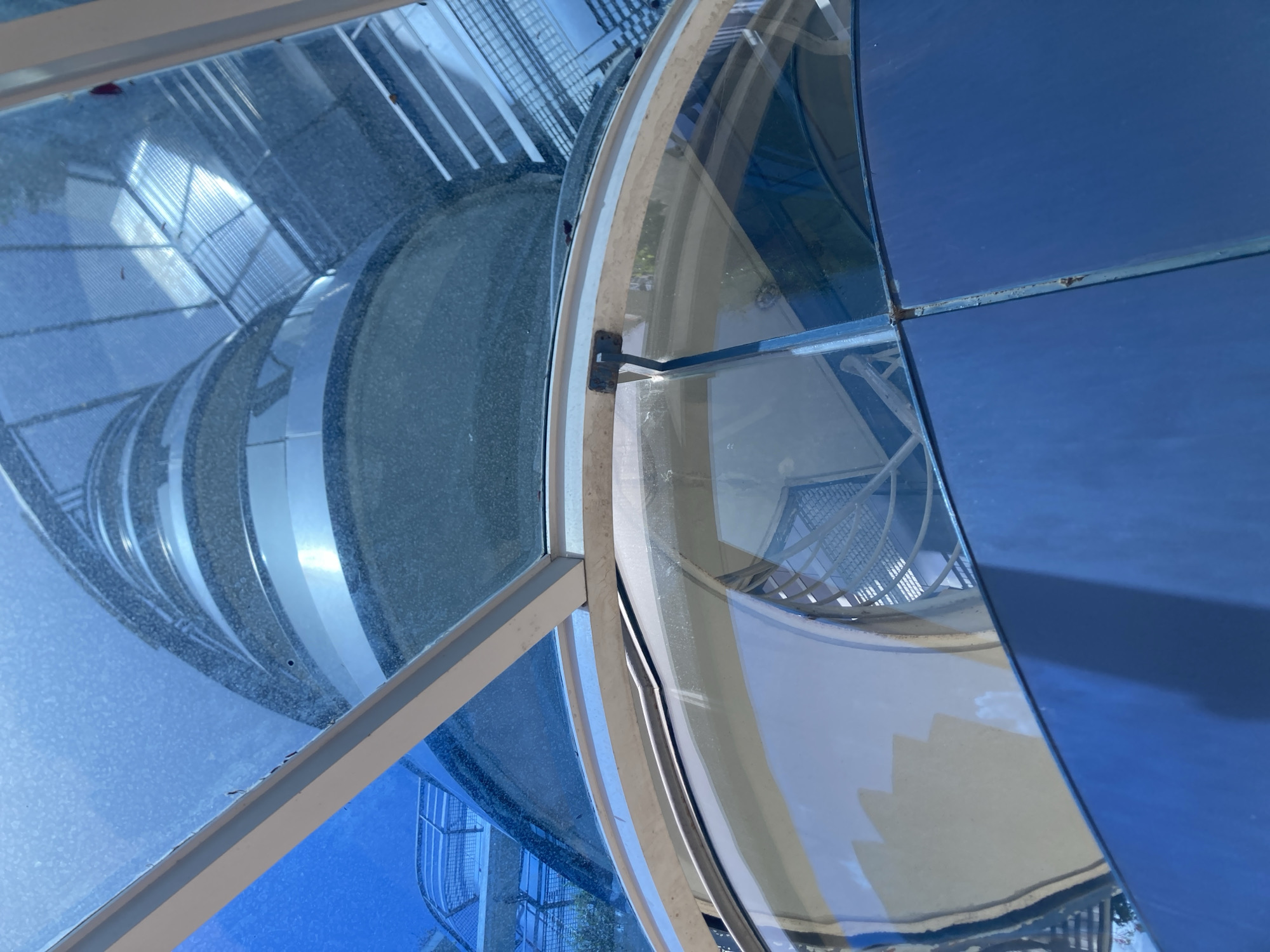 This is a low-angle, eye-level outdoor photograph looking up at the helical staircase of the Torre Agbar in Barcelona, Spain. The lighting is bright, suggesting it is daytime. The scene is dominated by shades of blue and metallic silver.
The foreground shows a curved, dark blue metal structure, possibly a wall or railing, with a vertical seam. Above this, a large curved glass panel is visible, reflecting some of the surroundings, including what appears to be a light-colored structure with angular shapes and a cloudy sky. A thin metal frame runs along the top edge of this glass panel, with a small, rusty metal hinge attached to it.
Beyond the foreground glass, the main subject is the exterior of the Torre Agbar. It features a spiraling structure with alternating bands of textured, dark material and smooth, reflective metallic silver rings. The texture of the dark material suggests it might be concrete or a similar rough-finished material, while the silver bands are highly polished and reflective.
Further up, the structure transitions to reveal glimpses of the interior, where a spiral staircase made of metal grating and white railings can be seen winding upwards. The overall impression is one of modern architecture with a distinctive helical design, viewed from an unconventional perspective that emphasizes the building's form and material contrast. The blue hue of the sky and potentially some translucent roofing elements overhead contributes to the dominant blue color palette of the image.