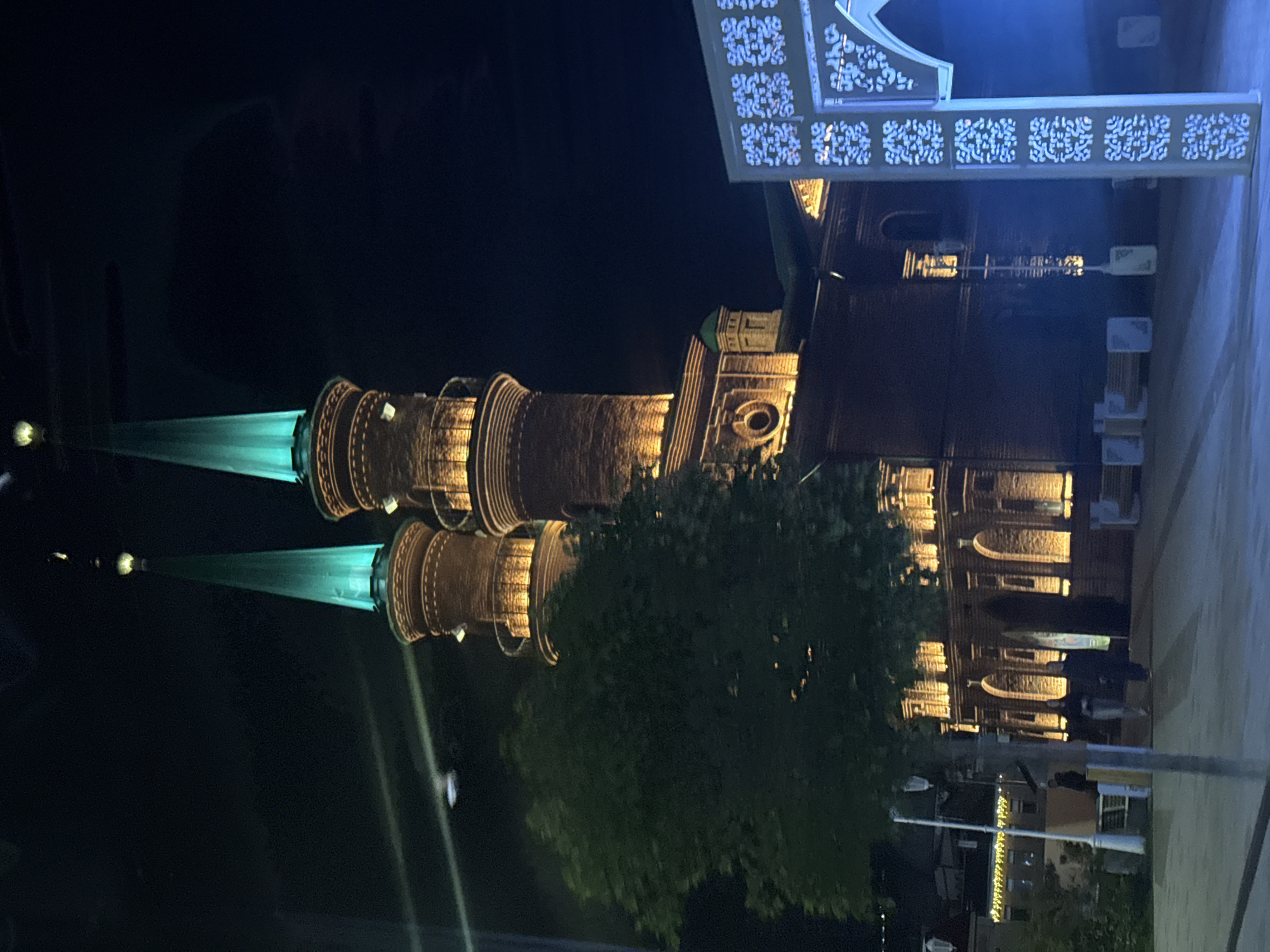 The image depicts the Mosque of the Hundred Names in Ufa, Russia, at night. The mosque features two prominent brick minarets, each topped with a pointed, teal-colored spire. The minarets are illuminated with warm, golden lights that highlight their architectural details. A large, dense green tree partially obscures the lower section of the minarets on the left side of the frame. In the foreground, to the right, a white decorative archway with intricate geometric patterns is visible, casting a blue light. Several people are present in the scene, blurred due to motion or distance, walking in front of the mosque's arched entrance. The background is dark, indicating it is nighttime, with some distant building lights visible on the left. The overall atmosphere is illuminated and festive, suggesting an event or a special occasion.